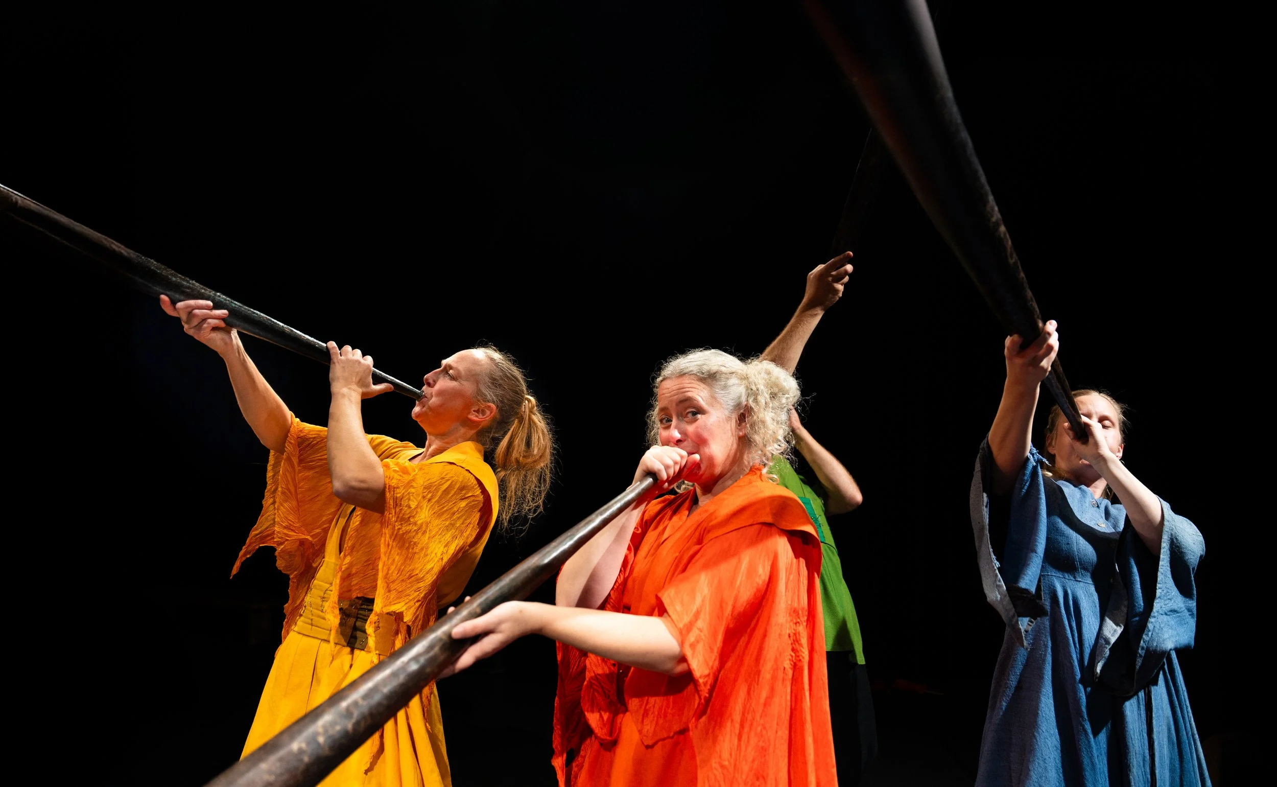 Tre kvinnor i färgglada kläder som spelar på långa instrument i mörkt rum.