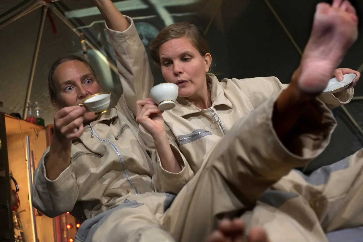 Två kvinnor i beige dräkter dricker från kaffeserviser och tittar förvånat på något.