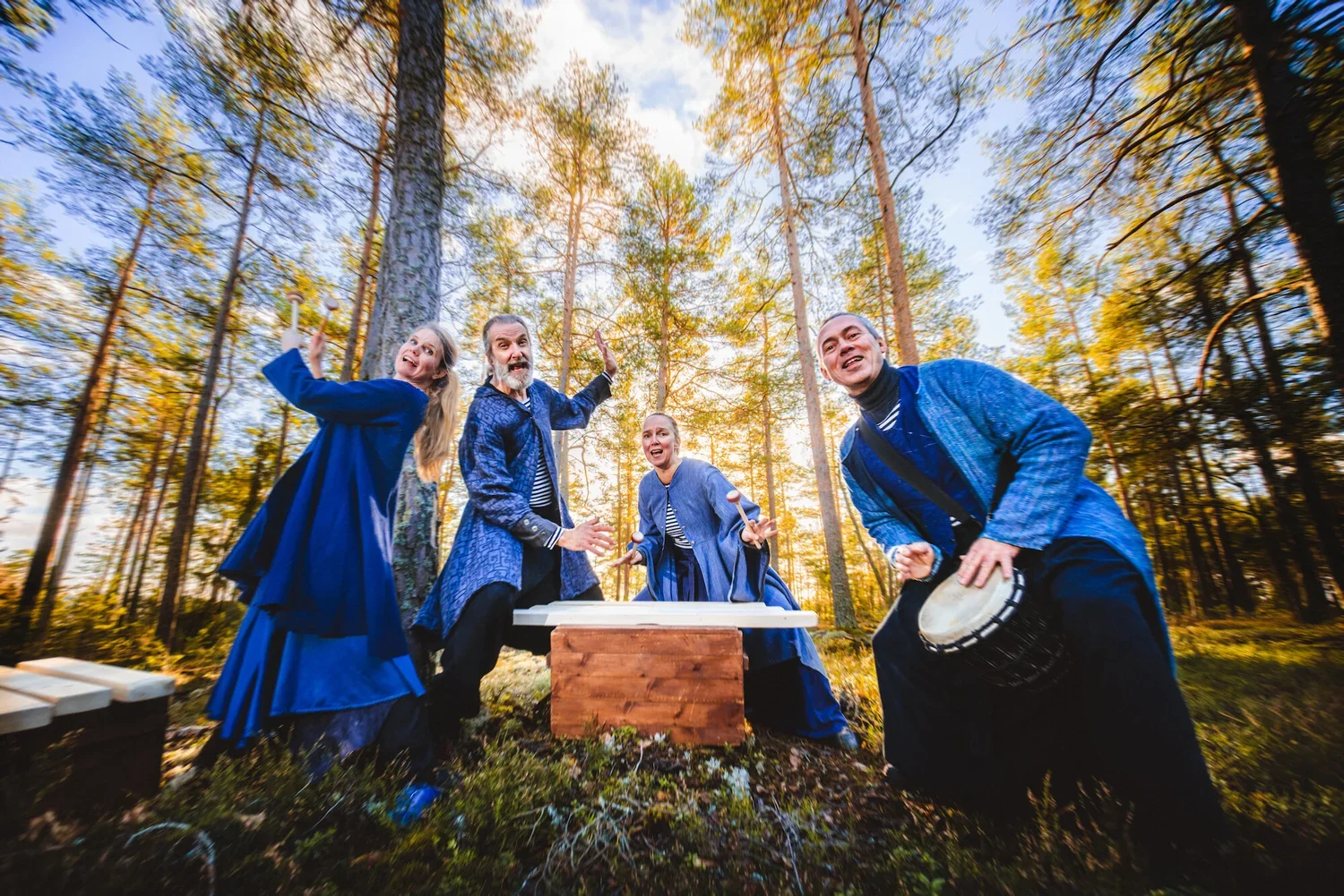 Grupp av fyra personer i blå kläder som sjunger och spelar instrument i en skog på hösten, med solen som skiner bakom träd.