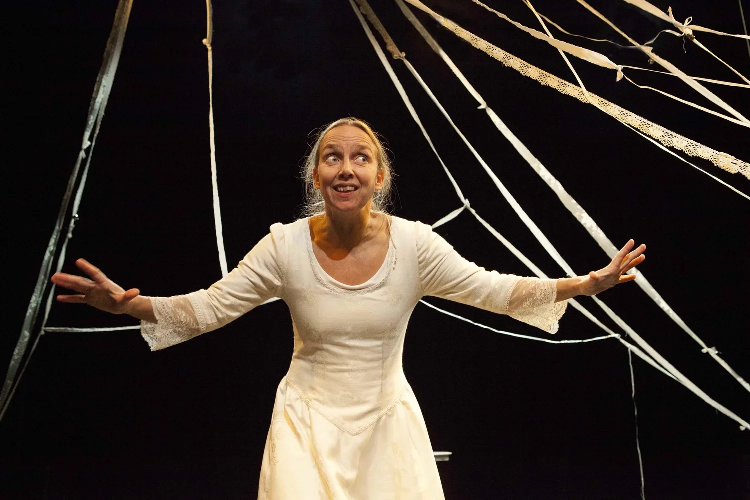 En kvinna i en ljus klänning framför en svart bakgrund med snörade linor, hon gestikulerar med armarna utsträckta.