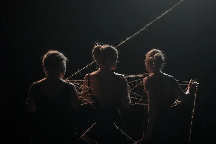 Tre personer i silhuett, omgivna av trådar i ett mörkt rum, med ett lätt ljus ovanifrån.