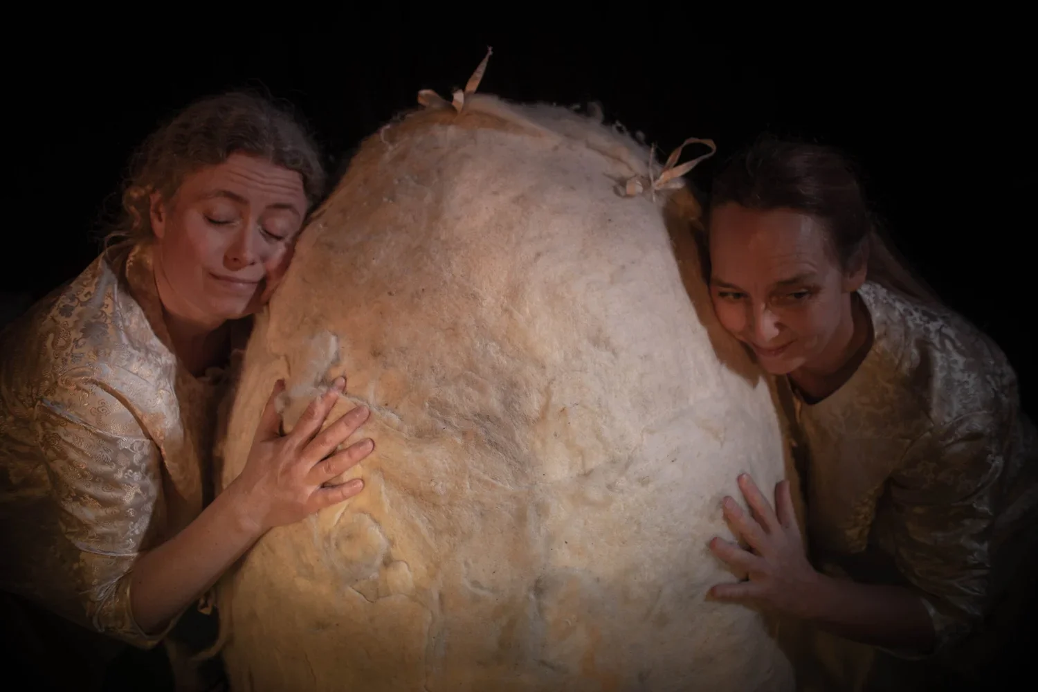 Två kvinnor i ljusa kläder omfamnar ett stort ägg av papier-maché, de ser känslosamma ut.
