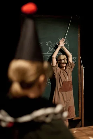 En person står framför en svängd tavla med ett kors på, i ett klassrum eller scenmiljö, och verkar prata eller presentera.