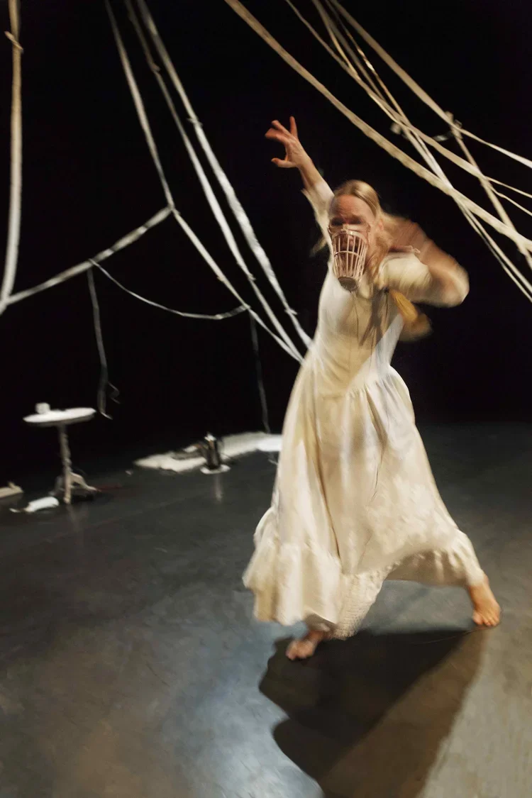 En person i en gammal klänning som dansar på en mörk scen med vita band hängandes runt omkring.