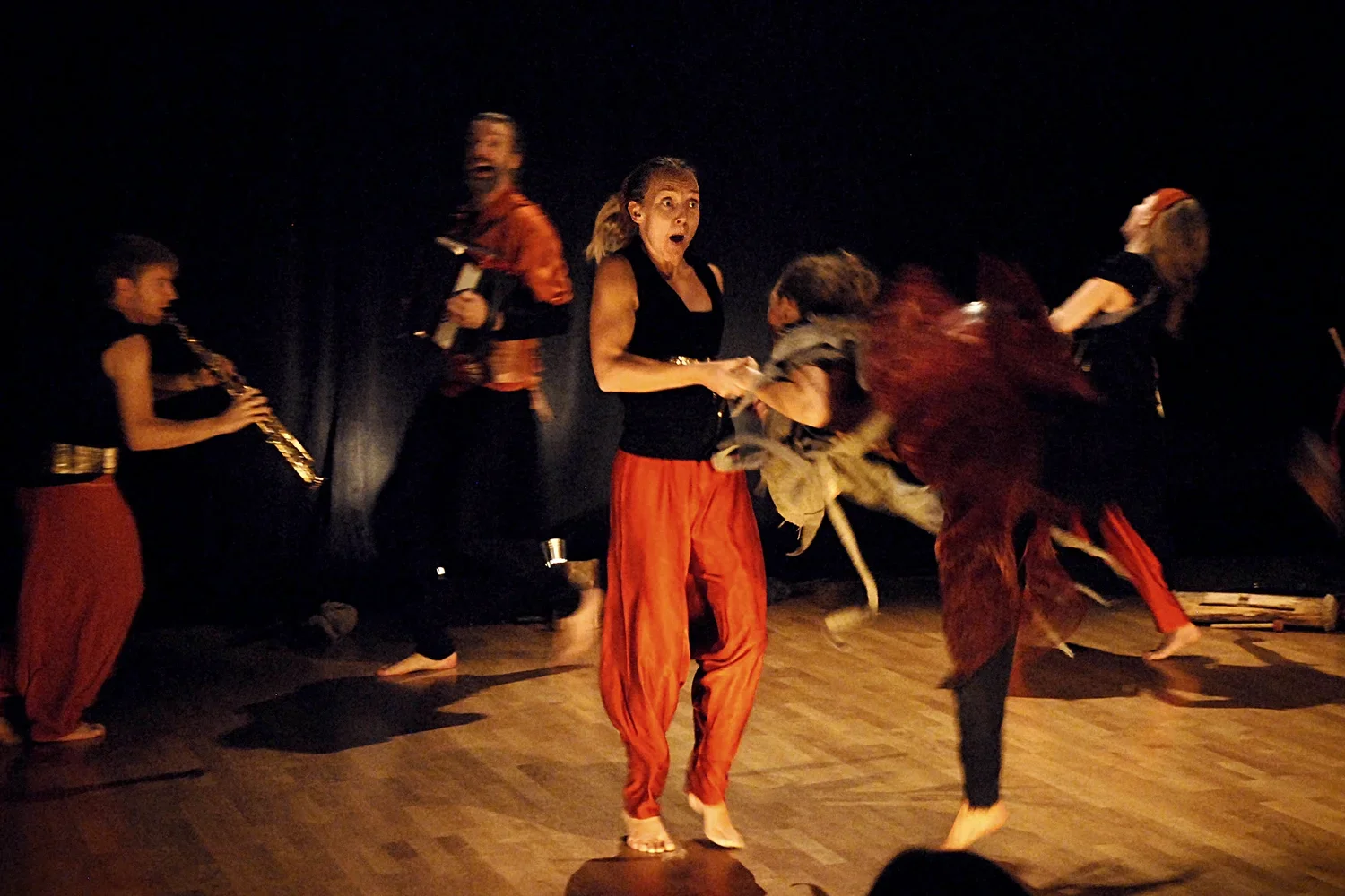 Flera personer som dansar och spelar instrument på scen, inklusive en kvinna i centralt fokus, med mörk bakgrund och trägolv.
