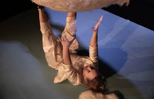 En kvinna i en beige klänning ligger på golvet och pekar uppåt, med ett blått och mörkgrått bakgrund.