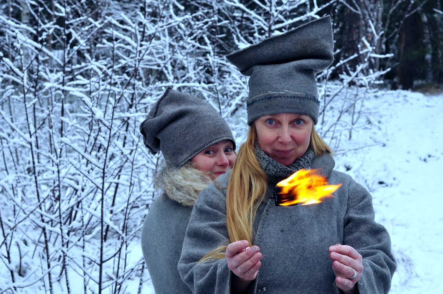 Två kvinnor i vinterkläder och mössor står i snöig skog, en av dem håller en liten eld.