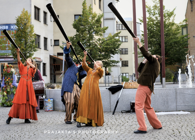 Fyra personer som spelar på långa pipor på en stadsgata, klädda i färgglada kläder, med träd och byggnader i bakgrunden.