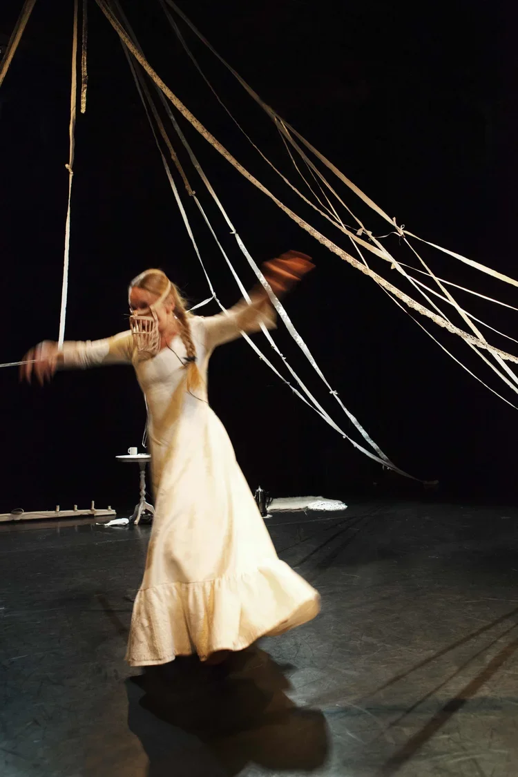 En kvinna i en lång, ljus klänning dansar på scenen med rop av tygband som sveper omkring henne, mot en svart bakgrund.