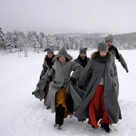 Grupp människor som går i snön, klädda i mörka pelsrockar med vinterhattar, omgiven av snötäckta träd och en snöig landskap.