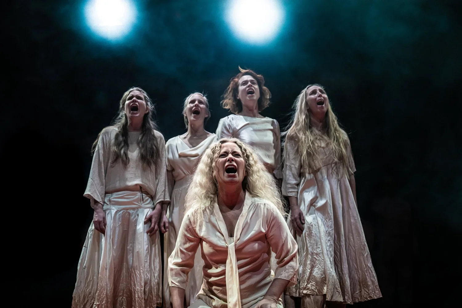Fem kvinnor på scenen som sjunger eller reciterar med öppna munnar i ett kraftfullt uttryck, klädda i ljusa kläder, under stark scenbelysning.