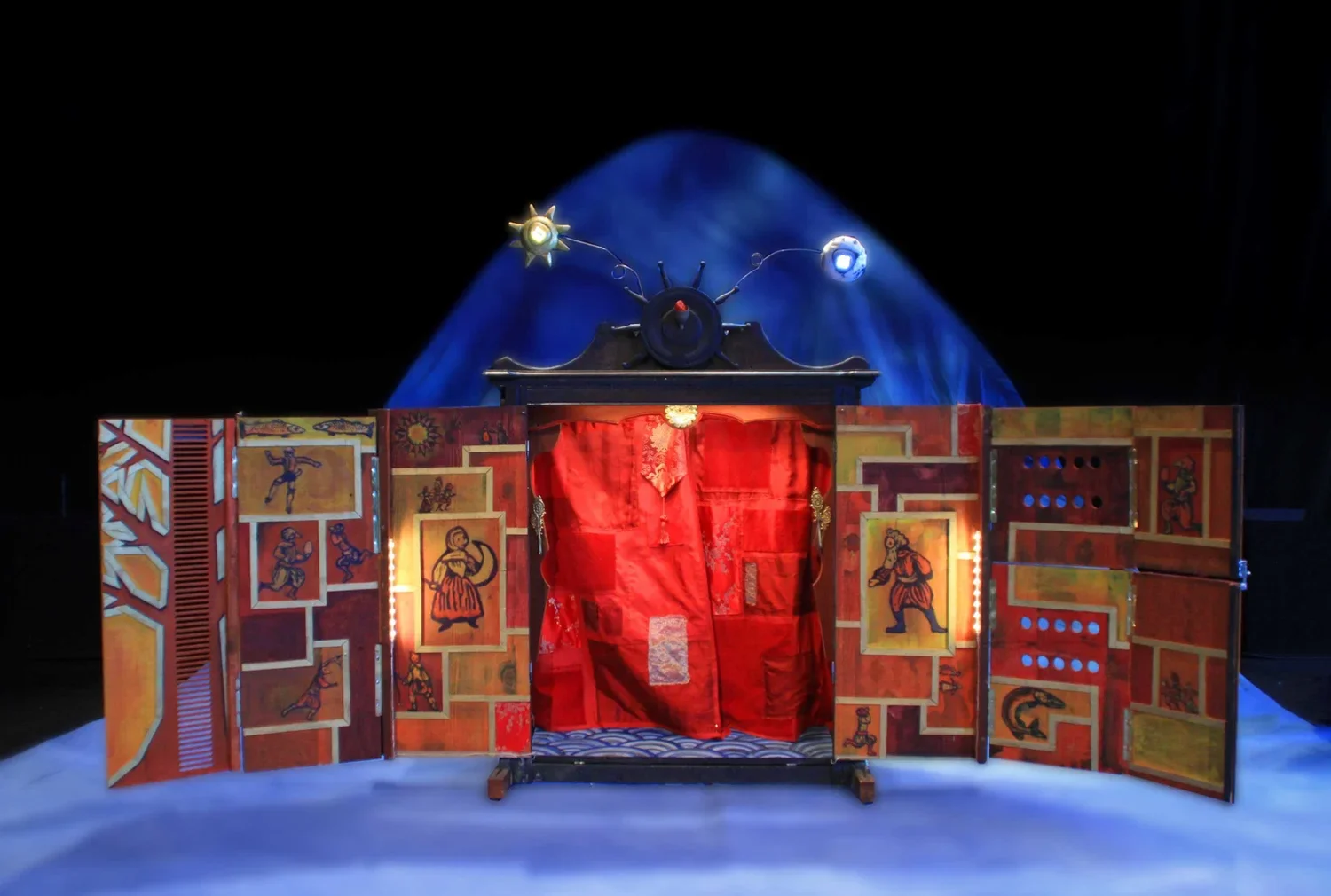 En teaterkuliss med röda gardiner, målade brickor och lampor. Bakom är en blå ljuspunkt mot en svart vägg. Tre konstnärliga ornament ovanpå, en som ser ut som en klocka med antenner som leder ut till små lampor.