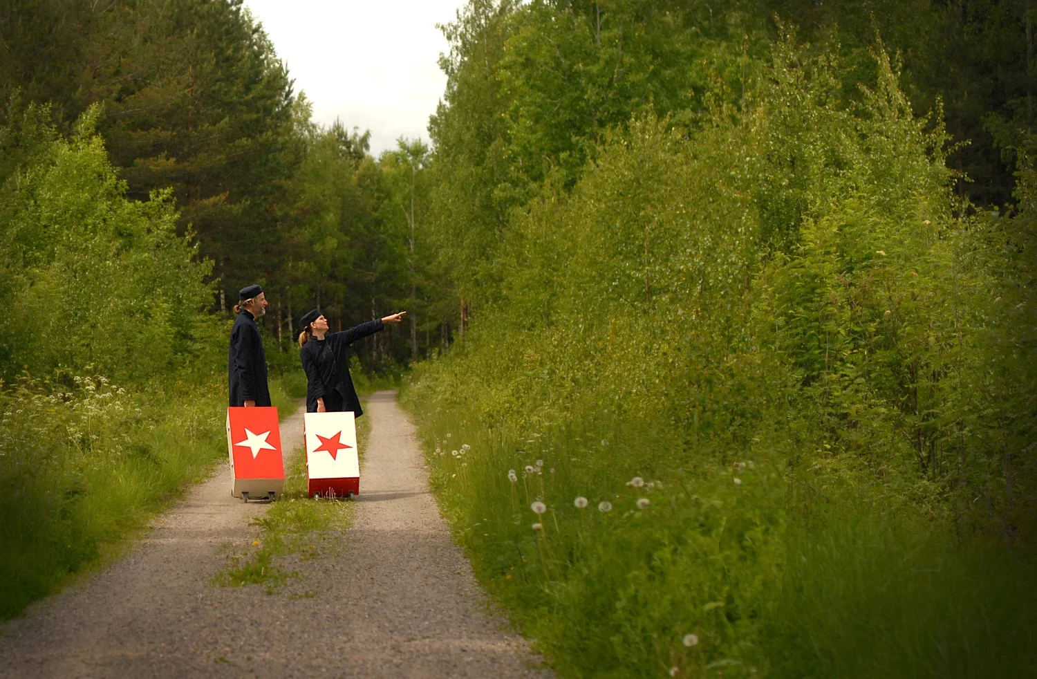 Två personer som bär svarta kläder står på en grusväg, den ena personen pekar mot något i skogen, båda har små vita och röda koffertar med liknande symboler
