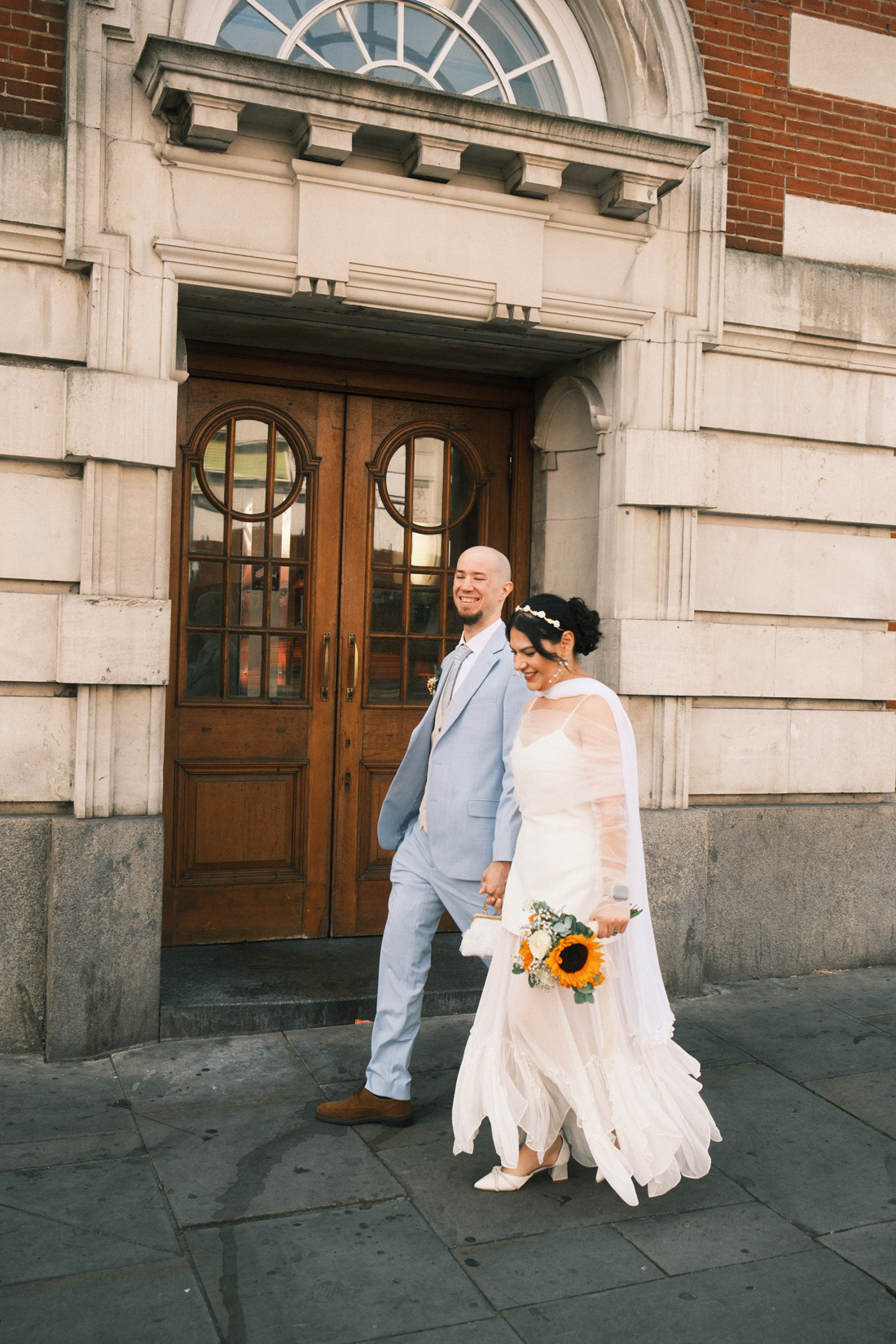 Una pareja de novios caminando afuera de un edificio con puertas de madera, la novia lleva un vestido blanco y sostiene un ramo con girasoles, el novio viste un saco azul claro y pantalones claros, ambos se ven felices.