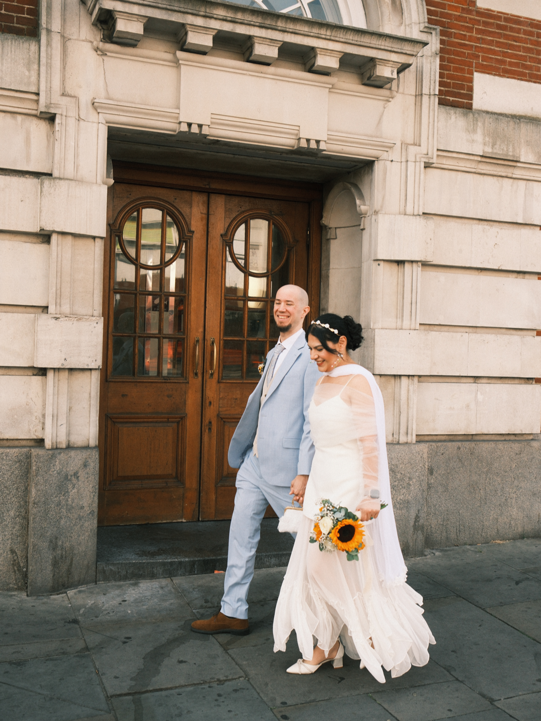 Pareja de recién casados caminando frente a un edificio de ladrillos y piedra, la mujer con vestido largo y bouquet de girasoles, el hombre con traje azul claro, ambos sonriendo.
