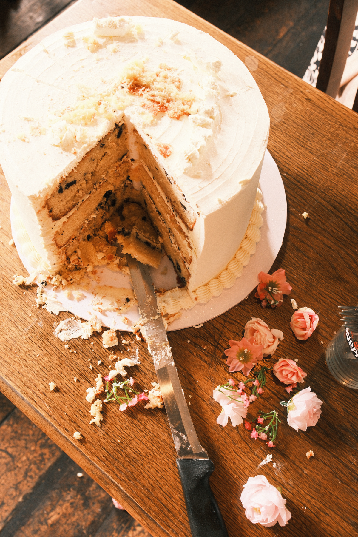 Pastel de chocolate con glaseado blanco y decoraciones de flores artificiales rosas y blancas en una mesa de madera, con una porción servida y restos de pastel en el plato y en la mesa.