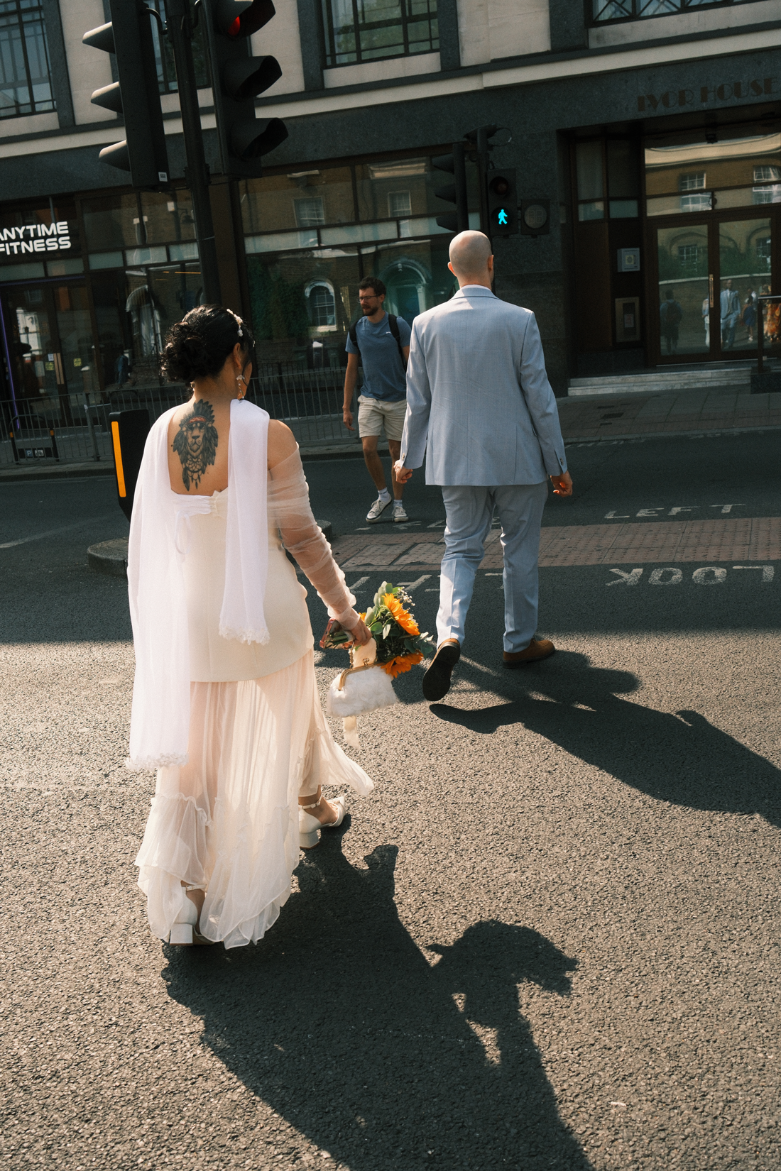 Una pareja vestida para una boda cruza la calle, con una mujer que lleva un vestido largo y sostiene un ramo de flores y un hombre con traje gris claro. En el fondo, un peatón con camiseta azul y pantalones cortos.