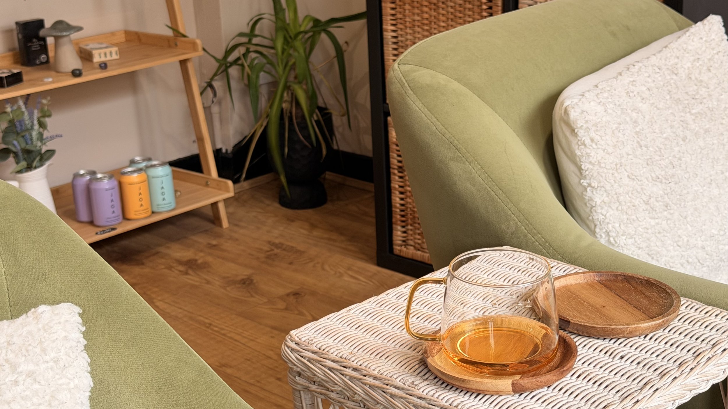 Close-up of a glass cup with orange beverage on a wicker table next to a wooden tray, with a cream-colored blanket, on a green chair.