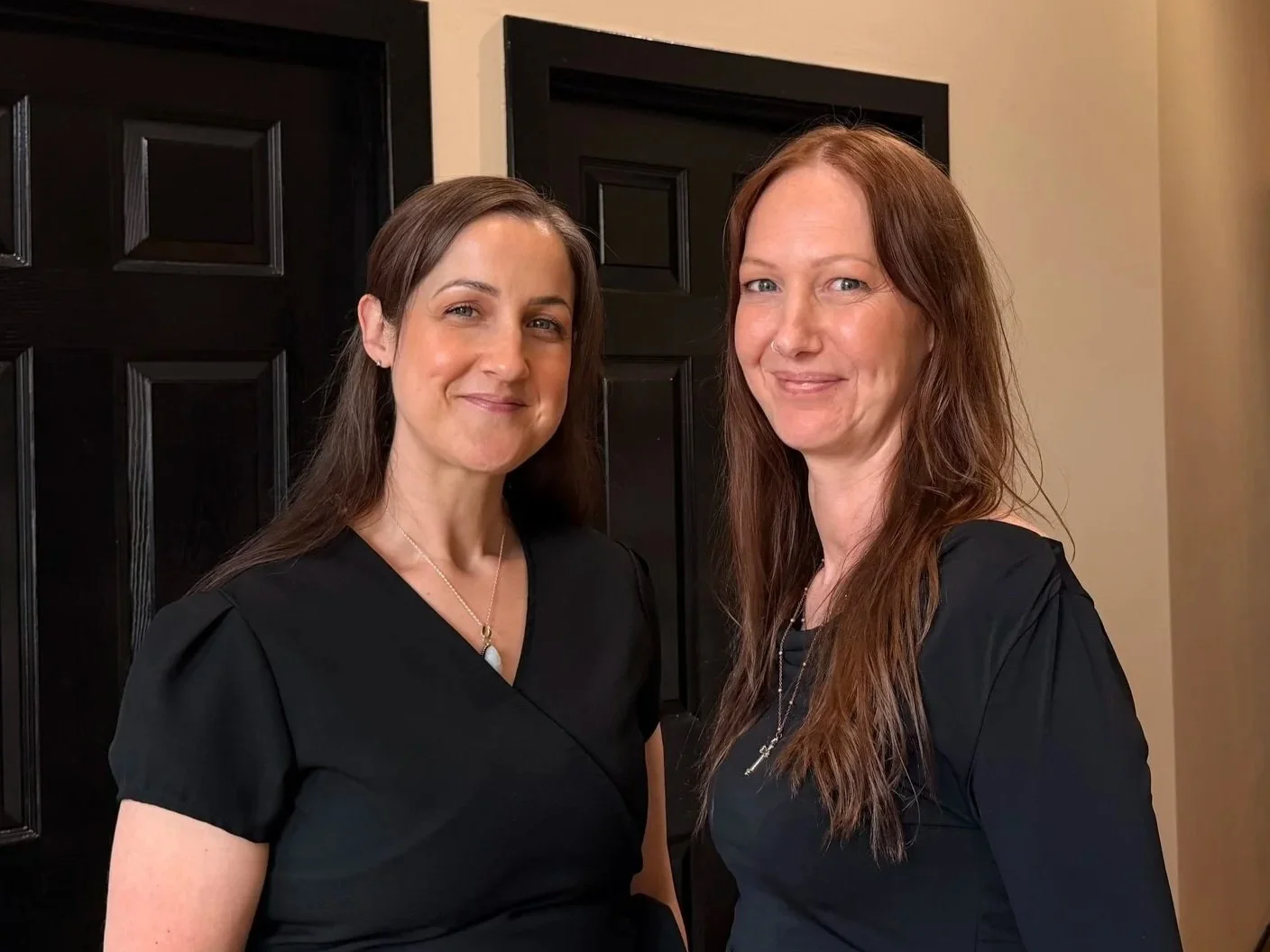 Two women with long hair, wearing black tops, standing indoors in front of a dark door and a beige wall, smiling at the camera.