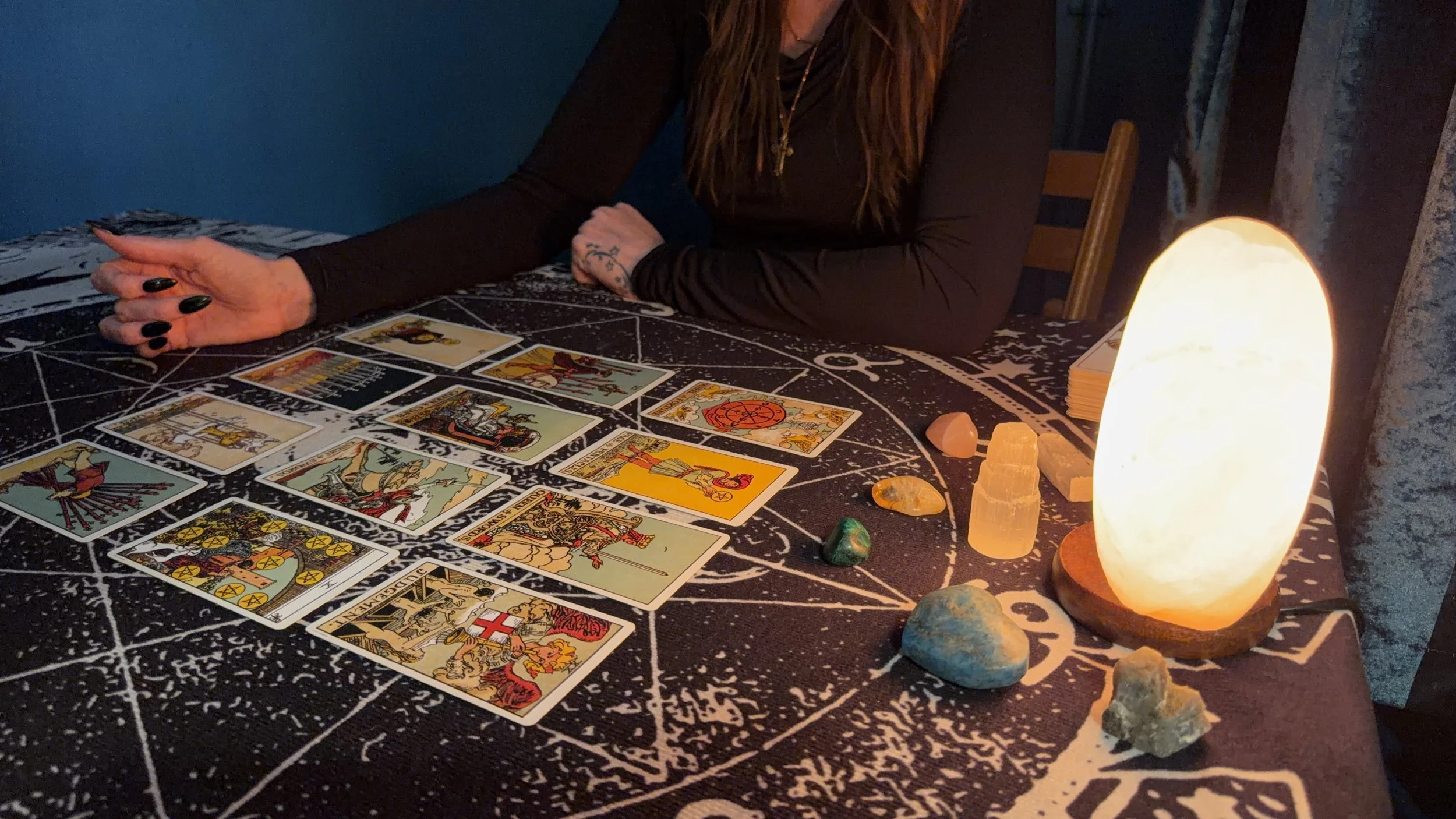 A tarot card reading setup with cards laid on a black cloth featuring white symbols, illuminated by a glowing salt lamp. A woman with long hair, wearing black and jewelry, sits with her arms crossed on the table, surrounded by colorful stones.