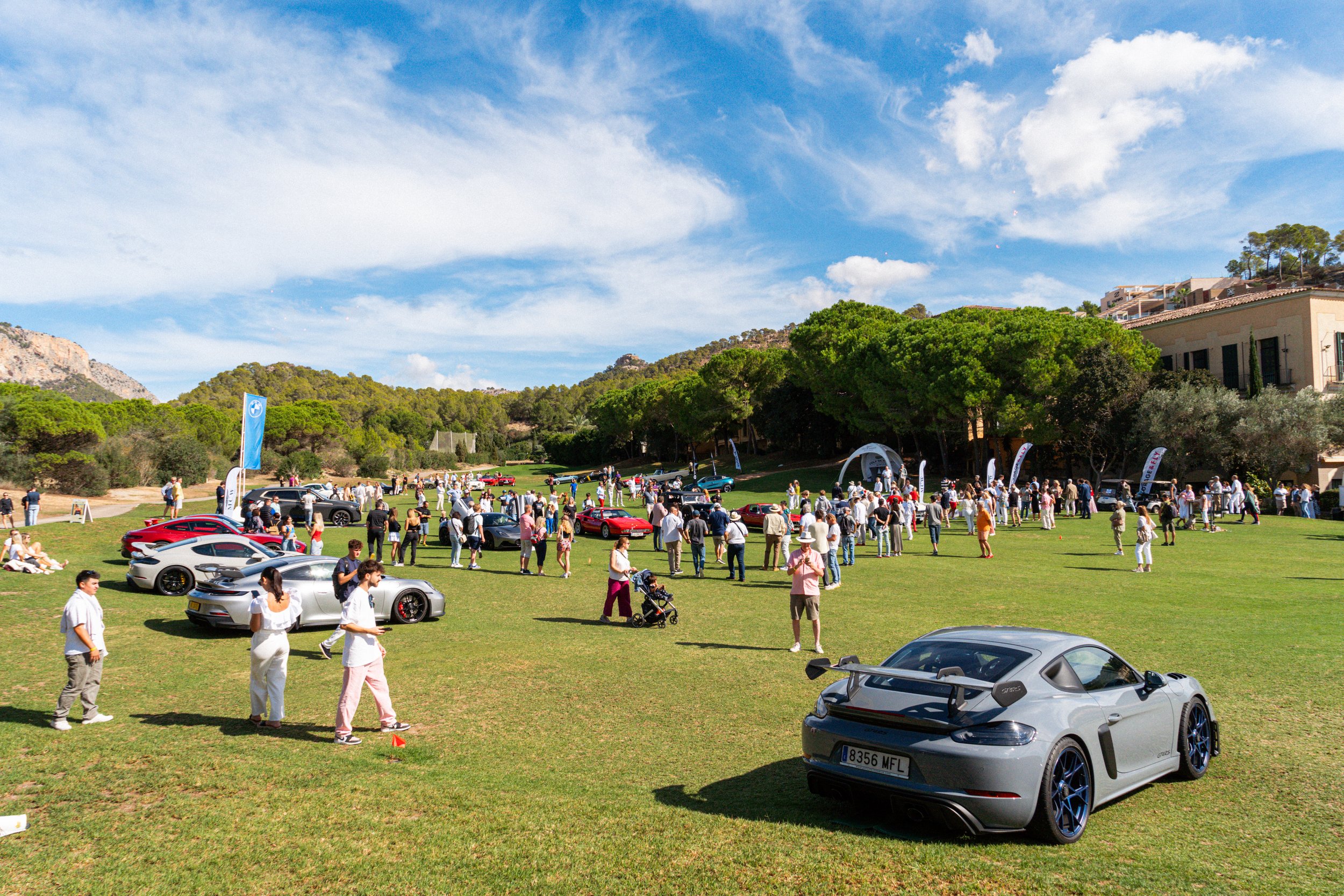 Exposición de autos deportivos en un parque en un día soleado con árboles y montañas en el fondo, muchas personas observando los autos y disfrutando del evento.