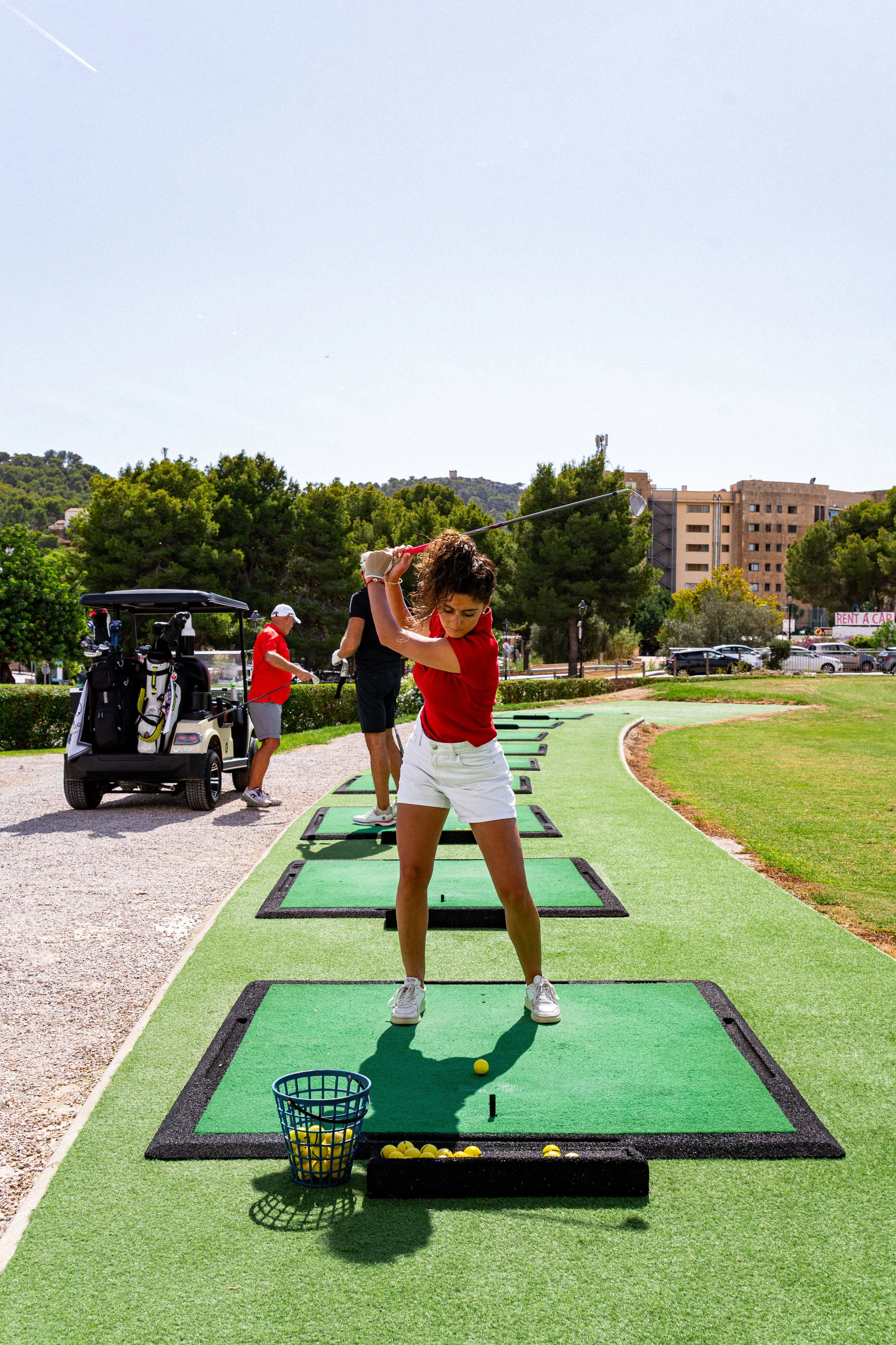 Mujer jugando golf en un campo al aire libre, con otros jugadores en el fondo y árboles verdes.
