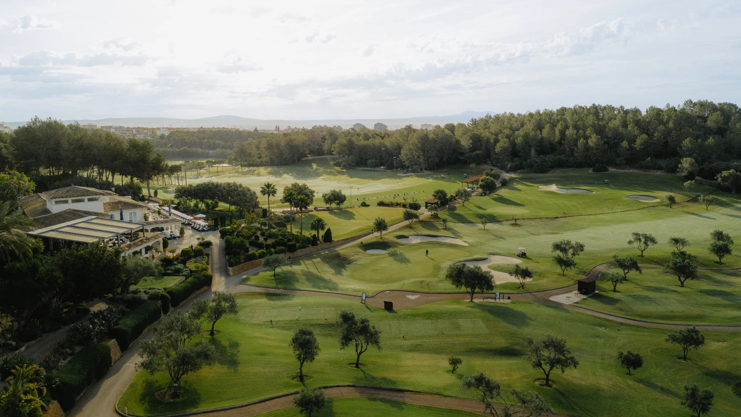 Campo de golf con césped asegurado, árboles dispersos, caminos y un edificio con terraza, rodeado de colinas y árboles en el fondo bajo cielos nublados.