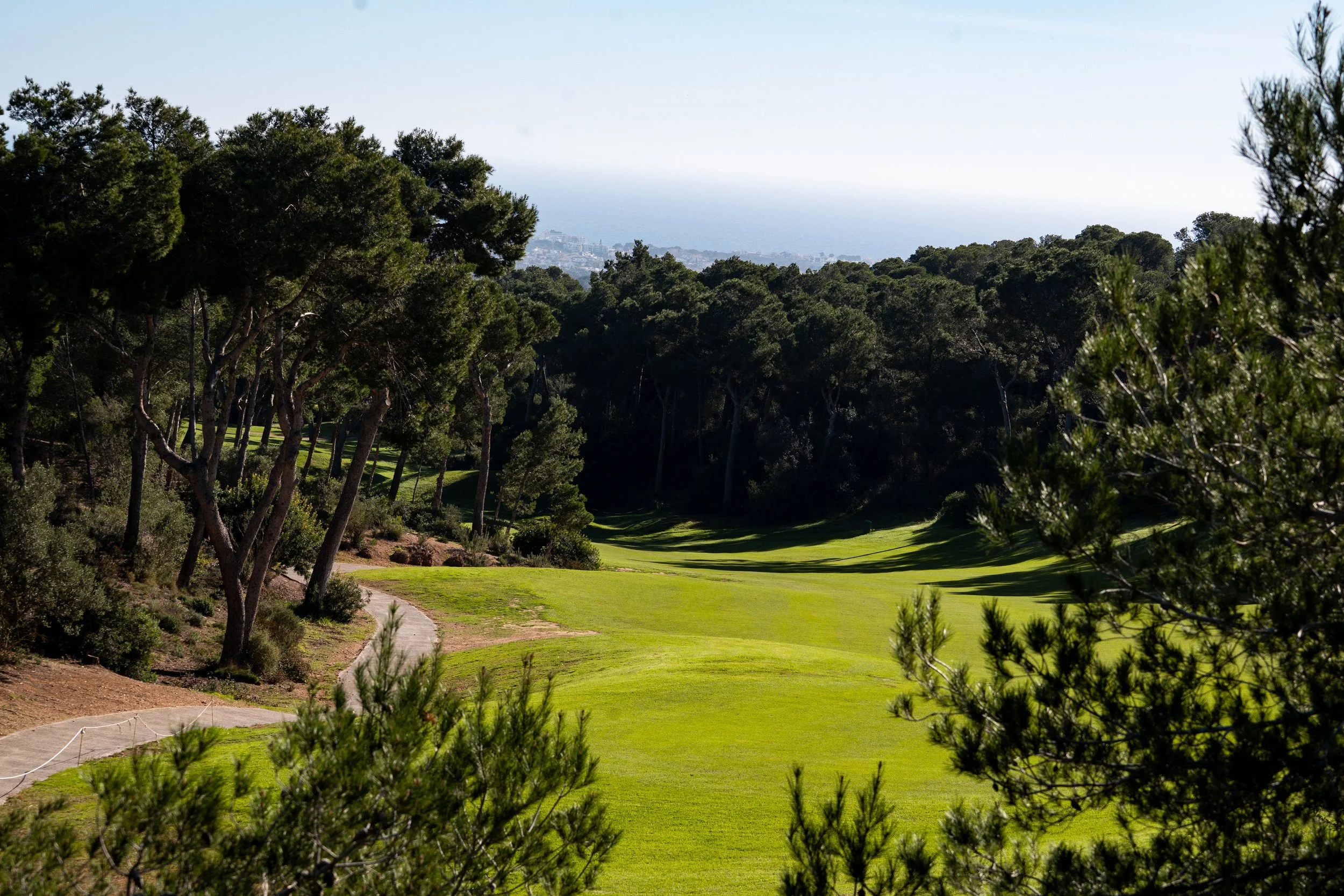 Campo de golf rodeado de árboles con vista al mar y ciudad en el fondo.