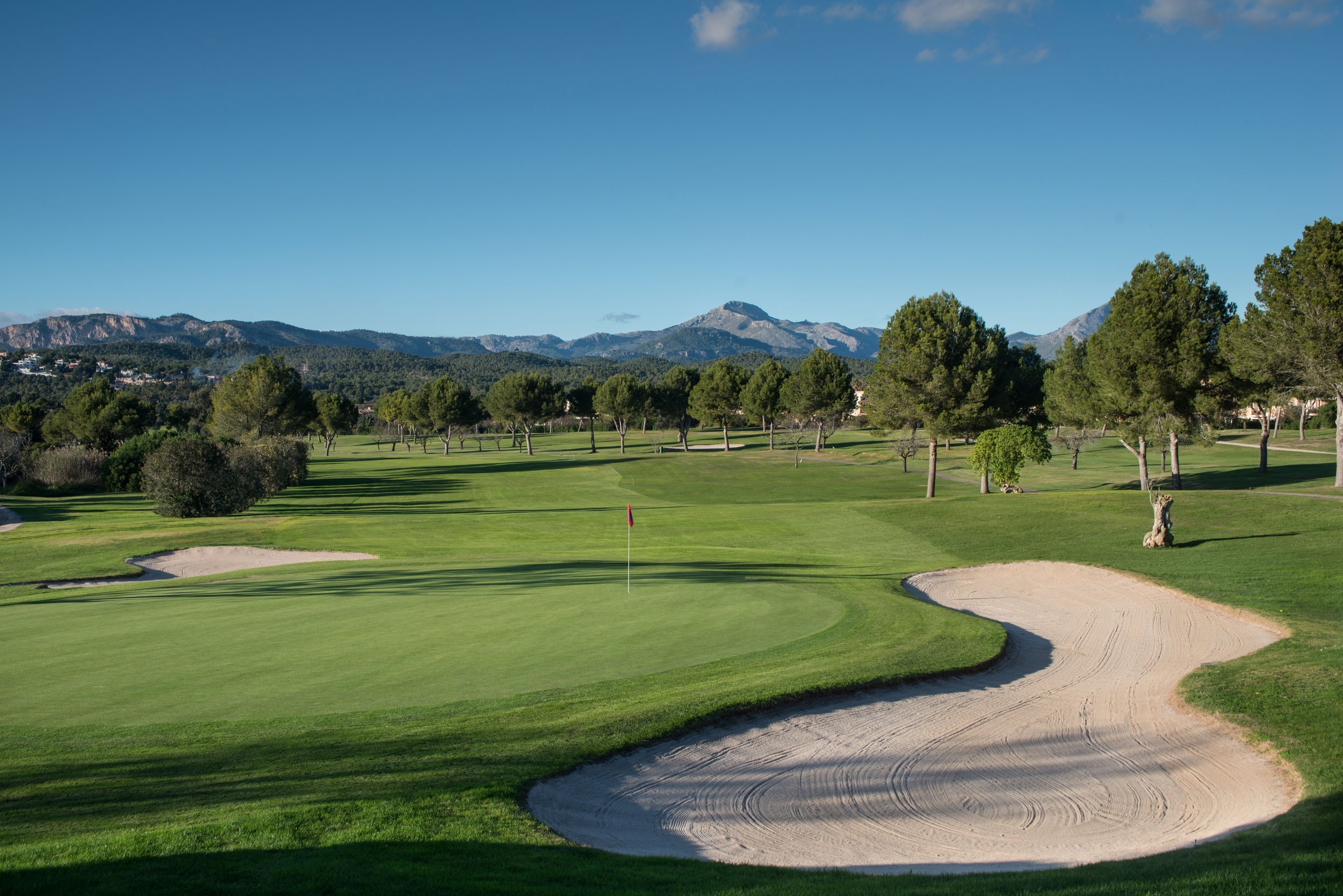 Vista de un campo de golf con césped verde, obstáculos de arena, árboles y montañas al fondo, cielo despejado con una pequeña nube.