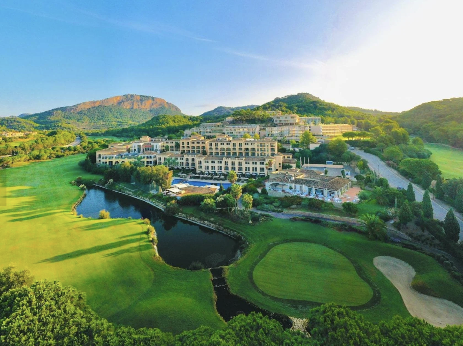 Vista aérea de un campo de golf junto a un complejo de edificios y montañas al fondo, con un río que atraviesa el terreno.