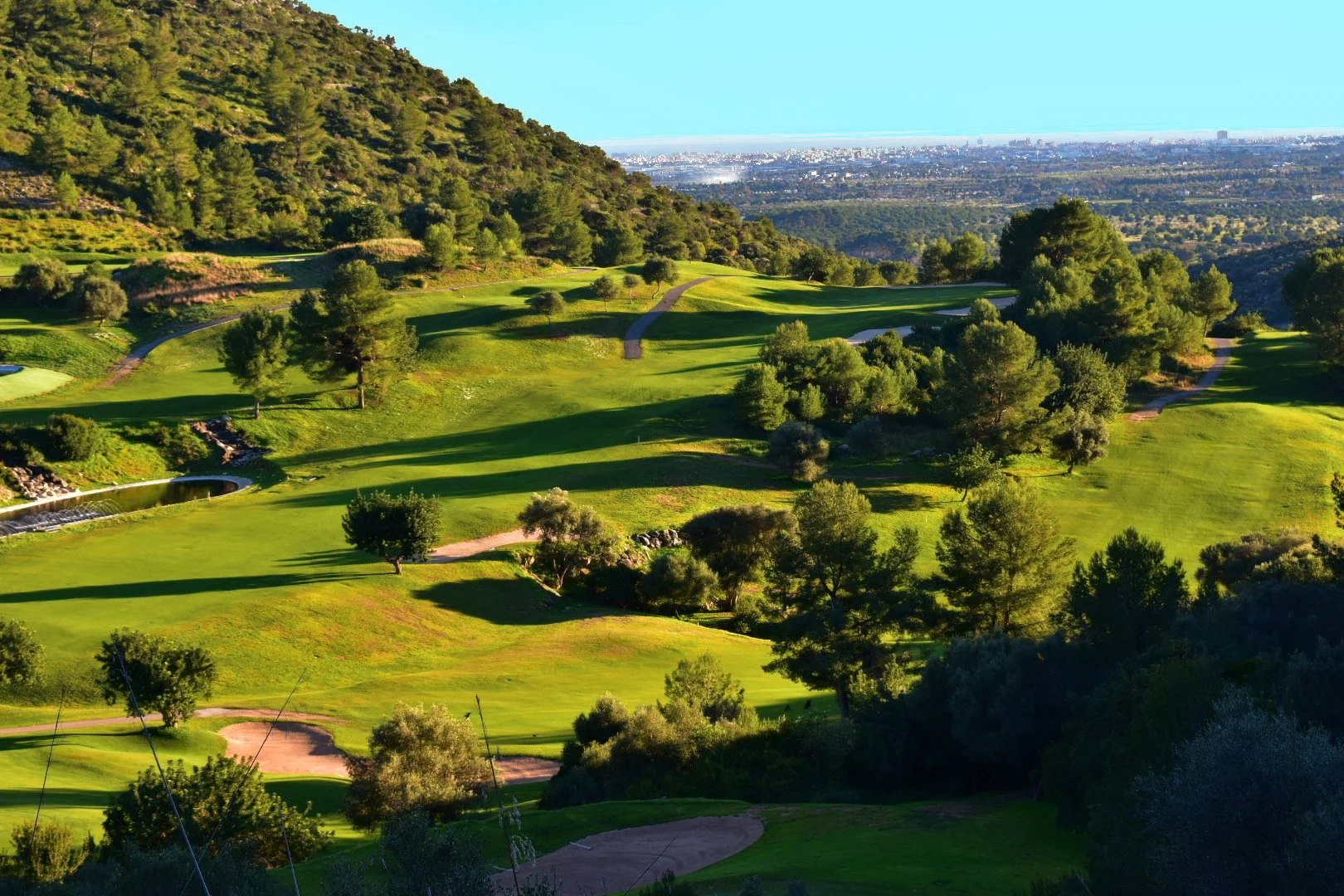 Un campo de golf rodeado de árboles y colinas verdes, con caminos y áreas de arena, en un día soleado con cielo despejado.