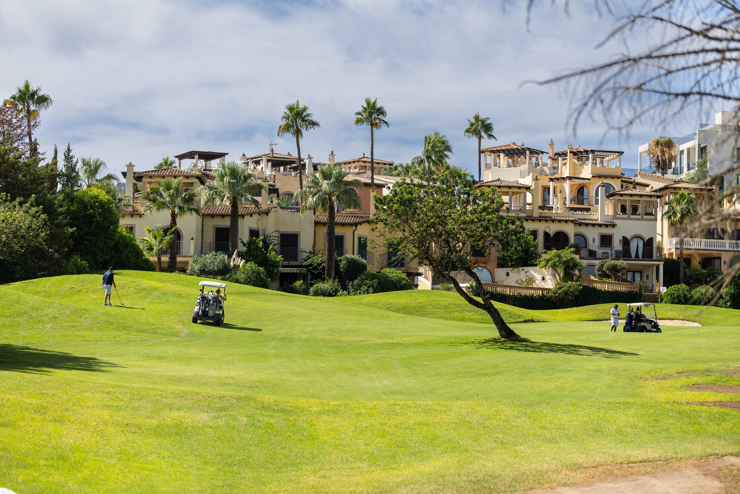 Campo de golf con varios jugadores y carritos, rodeado de árboles y casas de estilo mediterráneo.