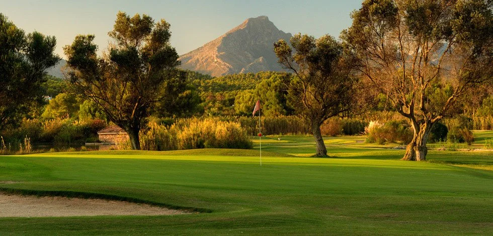 Curso de golf con árboles y montaña al fondo al atardecer.