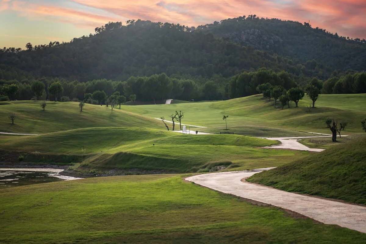 Paisaje de un campo de golf con caminos, árboles dispersos y colinas verdes, con montañas al fondo y un cielo con tonos rosados y naranjas al atardecer.