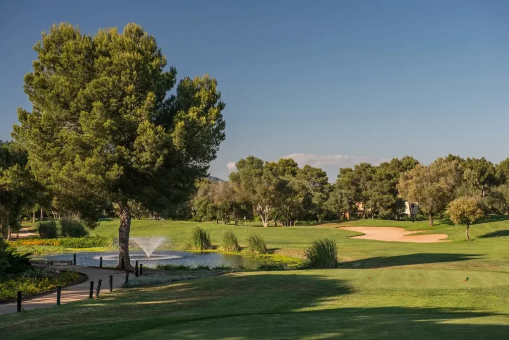 Un campo de golf con árboles, un estanque con fuente y arena de bunkers bajo un cielo despejado.