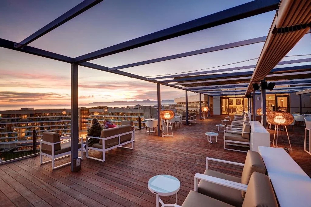 Terraza en la azotea con vistas al atardecer, muebles de patio blancos y sillones con cojines, lámparas de luz cálida, estructura metálica y techo de láminas, en una ciudad moderna.