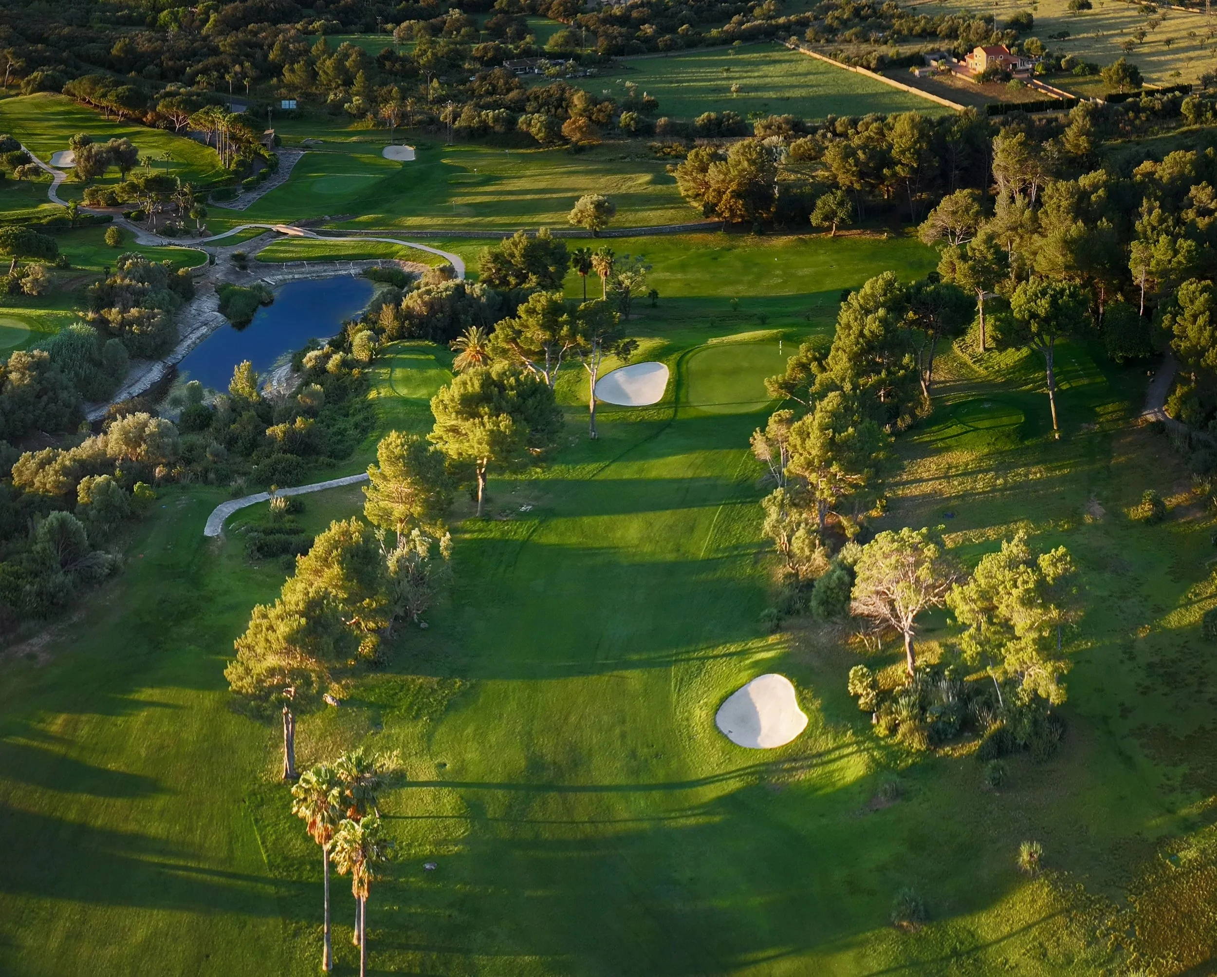 Paisaje aerial de un campo de golf rodeado de árboles y vegetación, con lagunas y caminos, en la hora del atardecer.