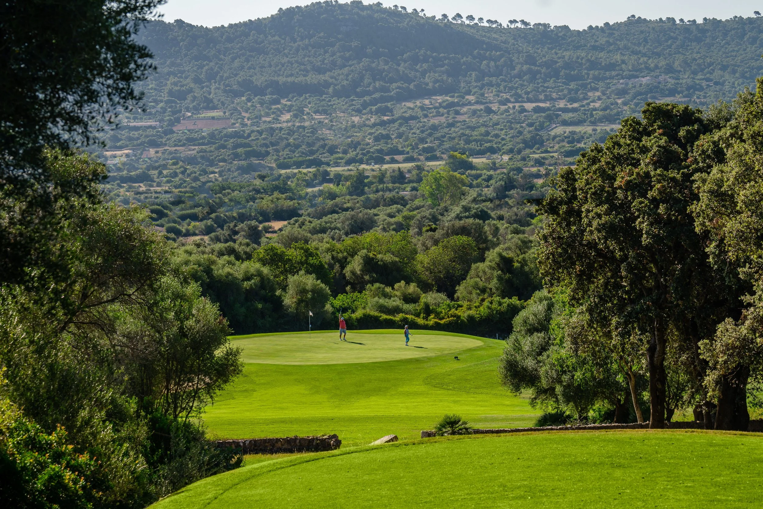 Campo de golf con montañas y árboles en el fondo, dos personas jugando golf en el césped verde.