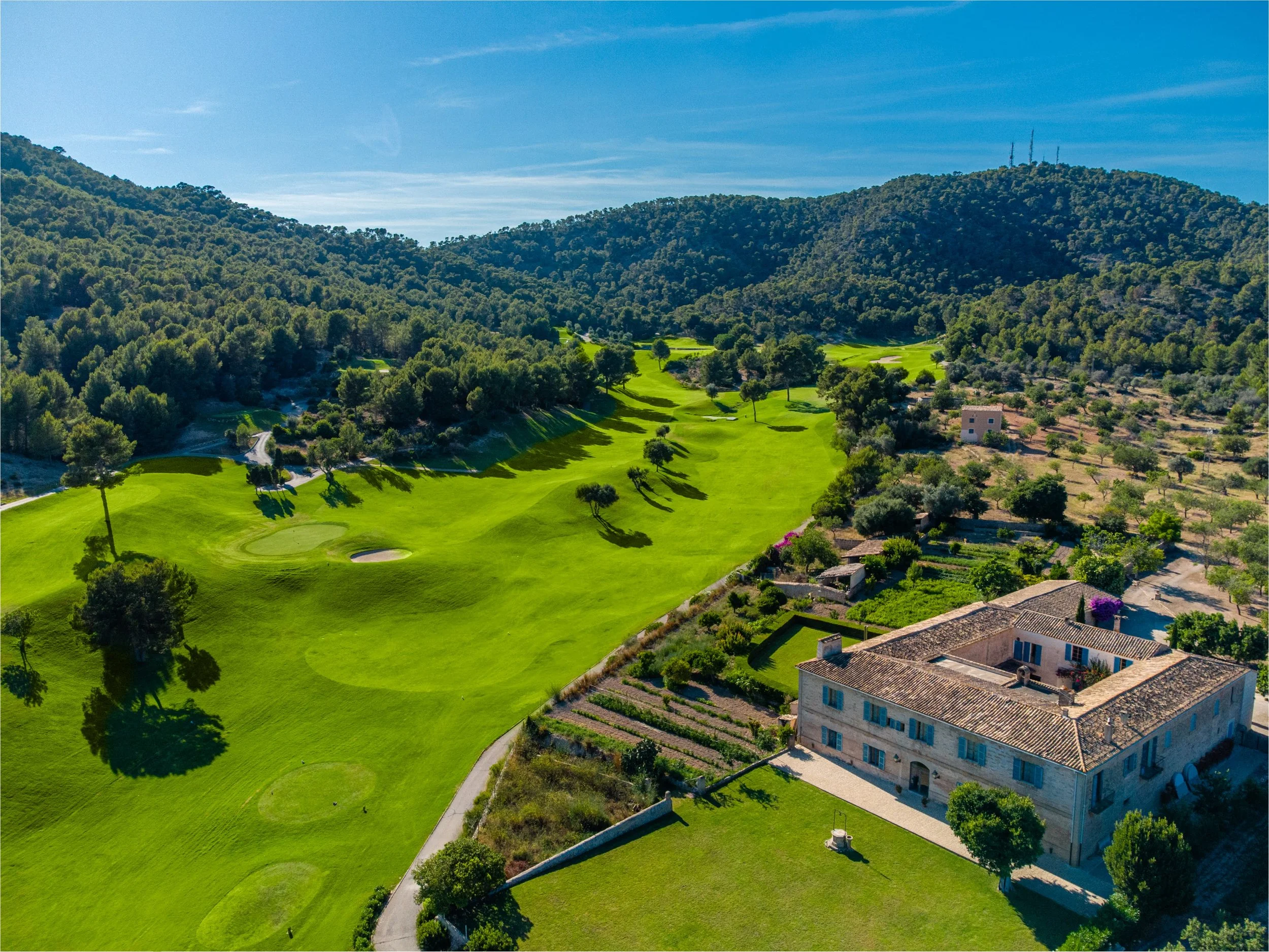 Campo de golf con césped bien cuidado, rodeado de árboles y colinas, con una casa grande de piedra en primer plano y montañas al fondo bajo un cielo azul con algunas nubes.