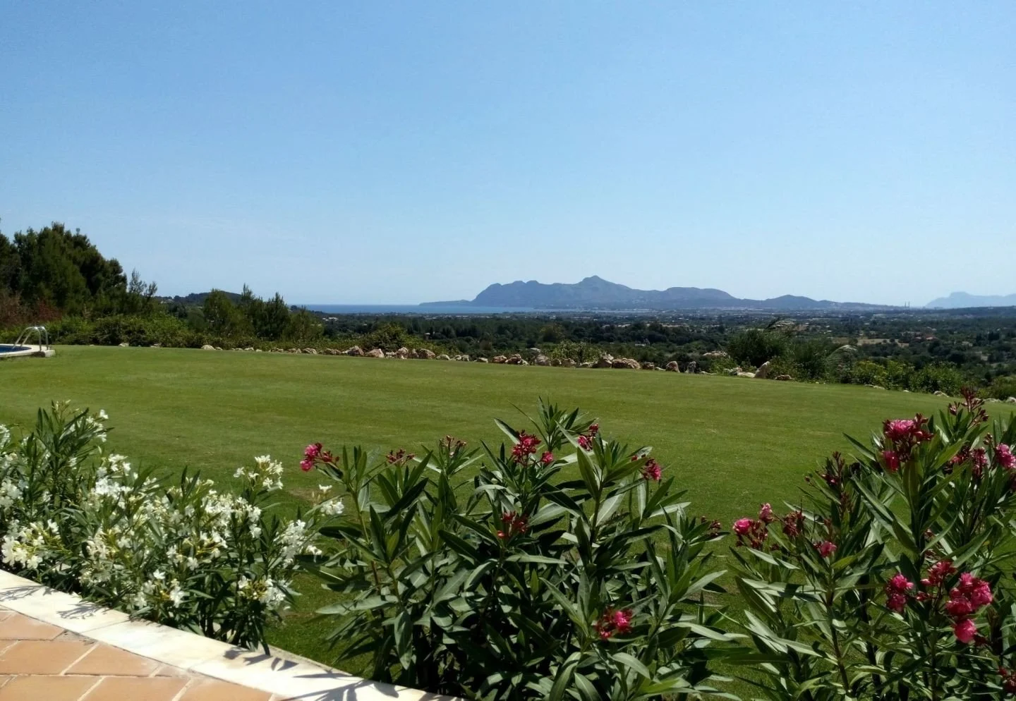 Vista panorámica de un jardín con césped verde, plantas con flores rosadas y blancas en primer plano, y montañas en la distancia bajo un cielo claro.