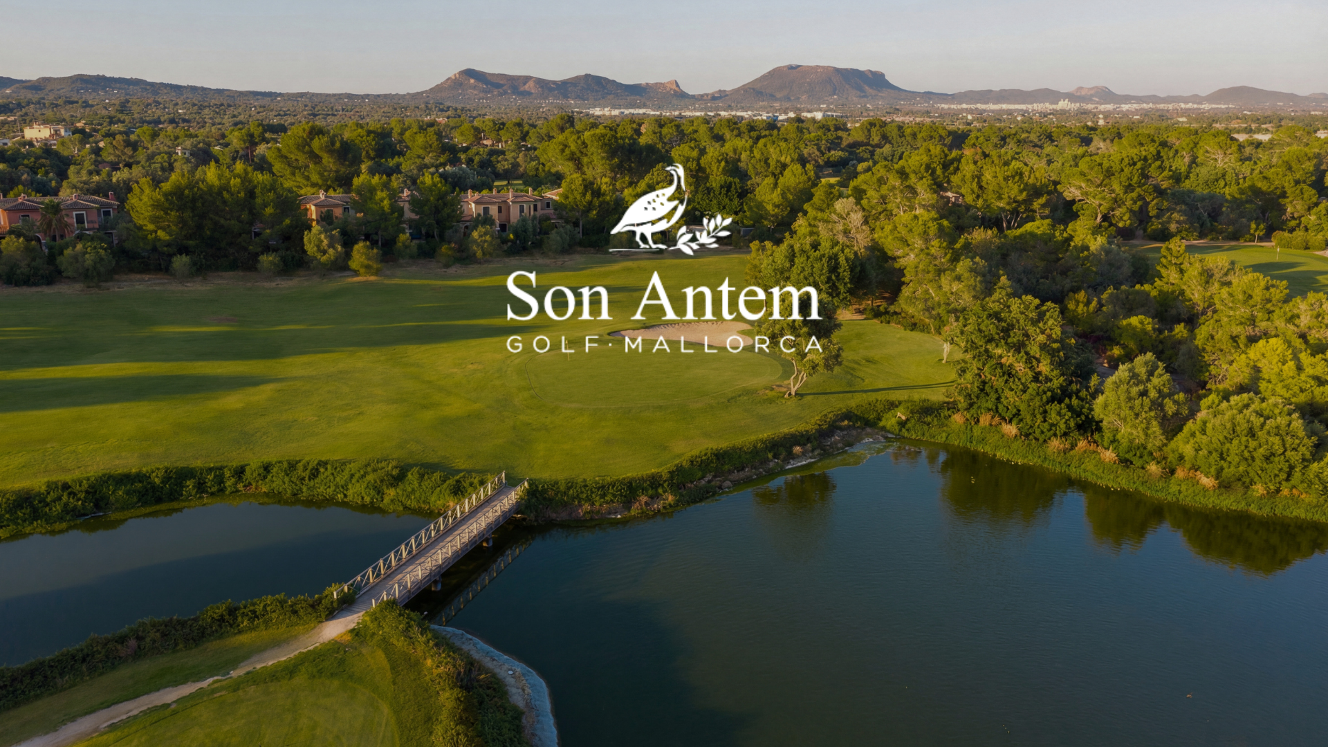 Vista aérea de un campo de golf en Son Antem, Mallorca, con laguna, árboles y montañas al fondo.