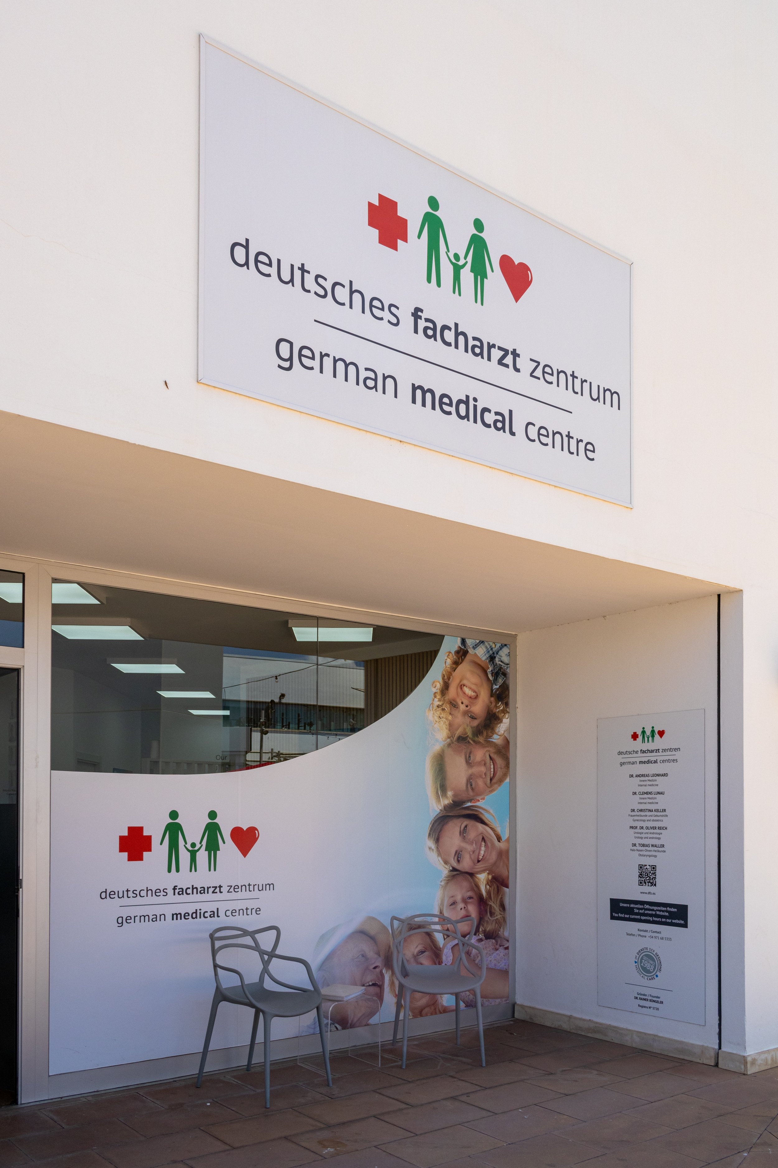 Cartel en un centro médico alemán con texto en alemán e inglés, incluyendo íconos de una cruz roja, una familia y un corazón, y varias fotografías de rostros sonriendo.