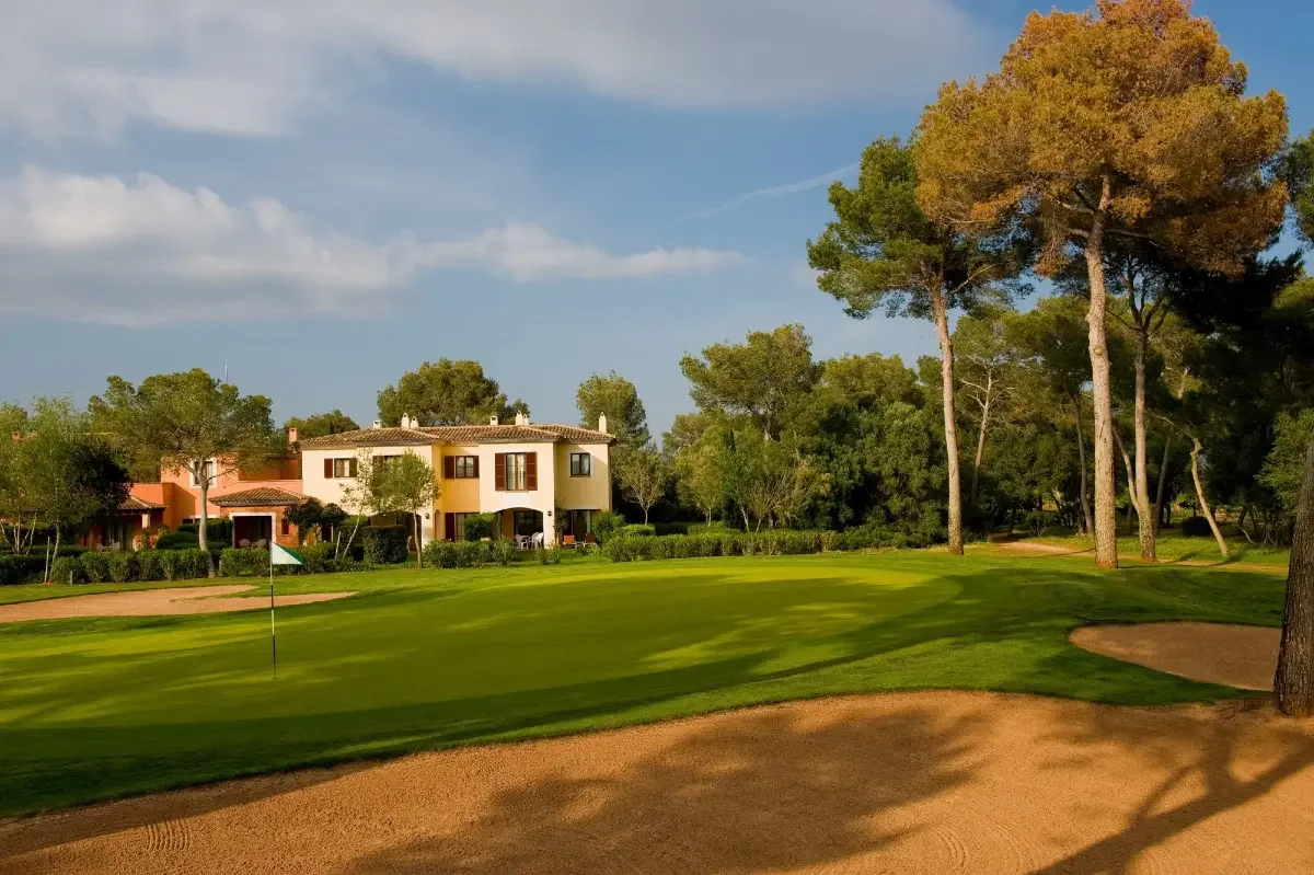 Campo de golf con casas residenciales y árboles en el fondo, cielo despejado con algunas nubes.