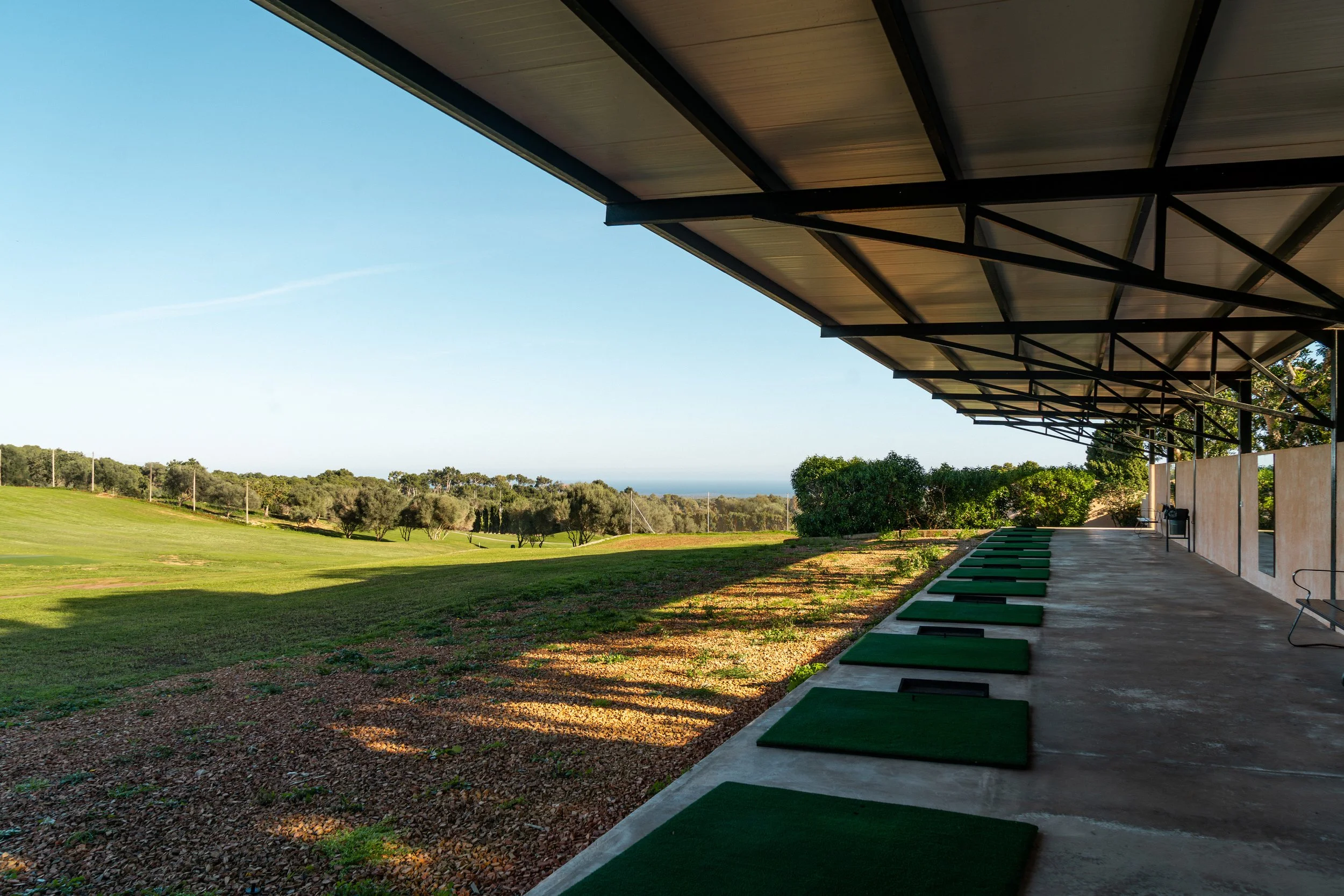 Área de prácticas de golf con varias alfombrillas verdes bajo un cobertizo y un campo abierto con árboles y cielo despejado al fondo.