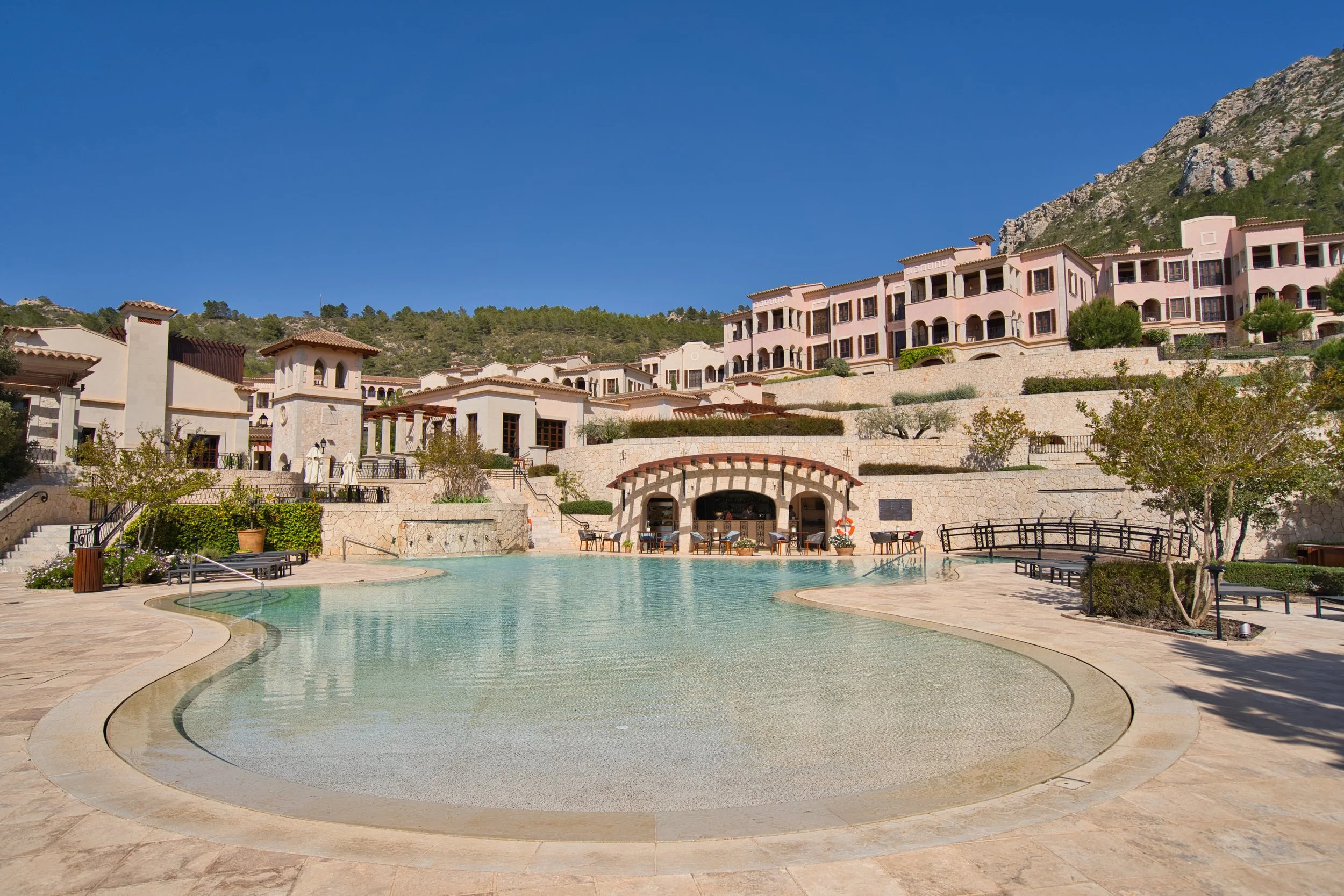 Piscina de forma curva con área para descanso y sombra, en un complejo residencial con edificios de estilo mediterráneo en las colinas, bajo un cielo azul.