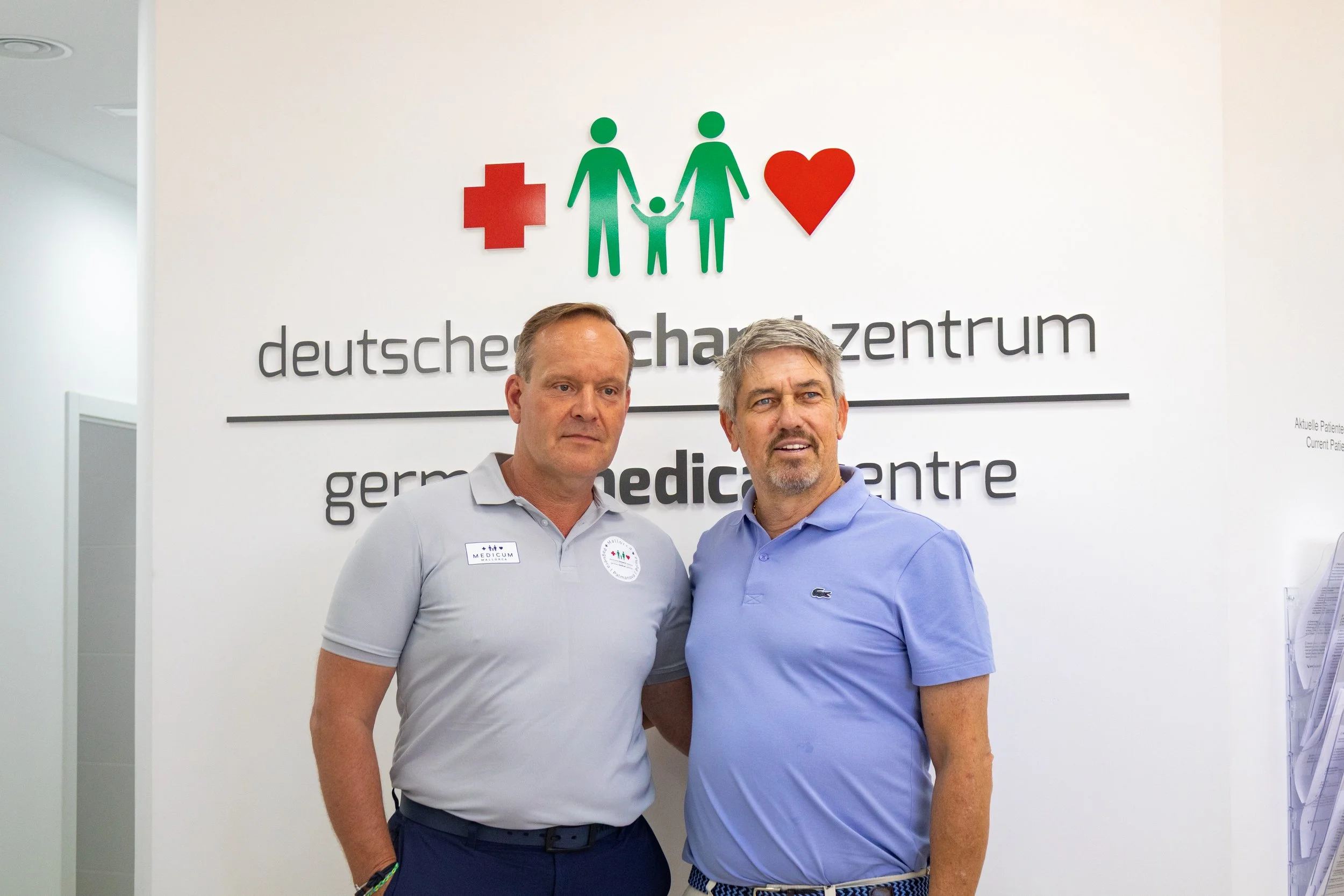 Dos hombres de pie frente a un cartel que dice 'deutsche gynäkologische zentrum' en un centro médico, con iconos de una cruz roja, una familia y un corazón.
