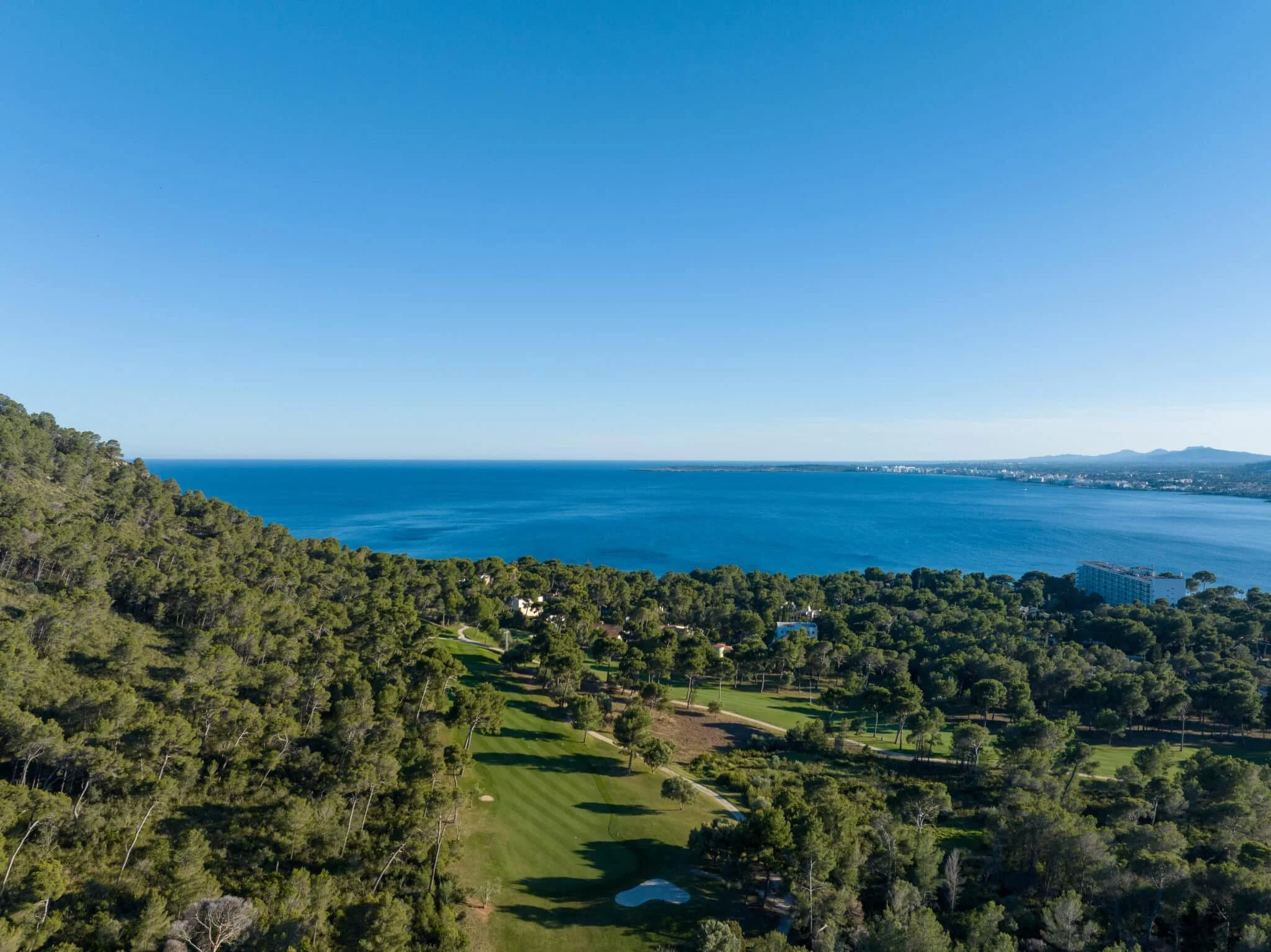 Vista aérea de un campo de golf rodeado de árboles verdes, con el mar y el horizonte en el fondo bajo un cielo despejado