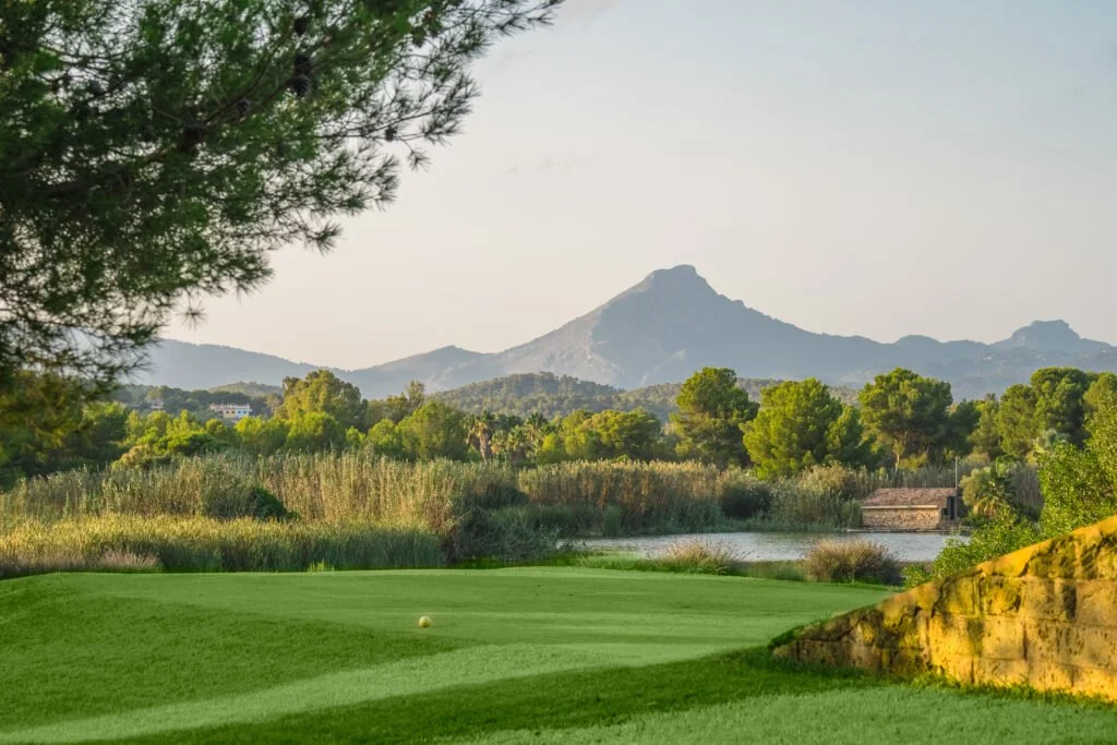 Vista de un campo de golf con césped bien cuidado, rodeado de arbustos y árboles, con montañas al fondo y un cuerpo de agua cercano.