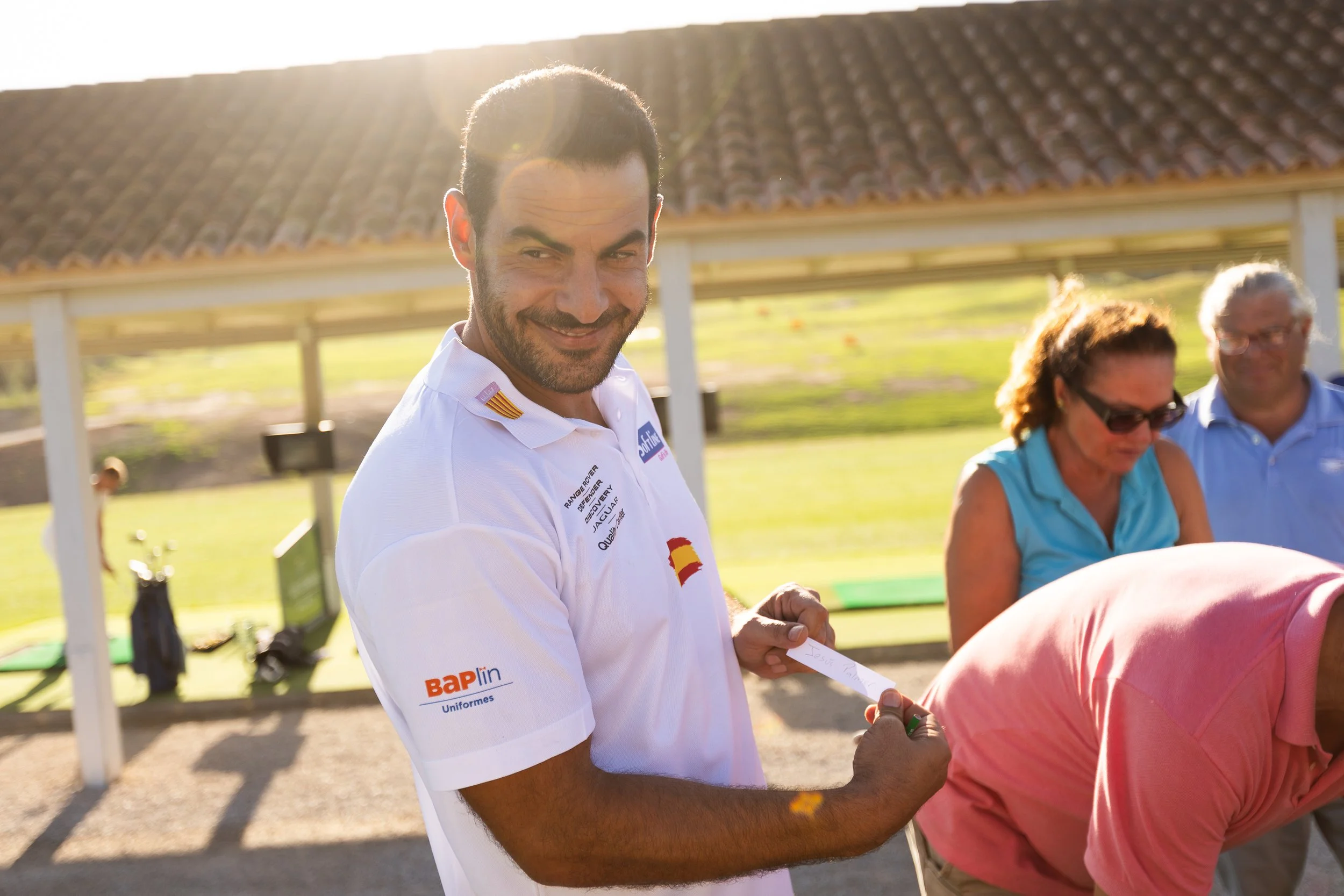 Hombre sonriendo sosteniendo una nota en un evento al aire libre con varias personas en el fondo, en un día soleado