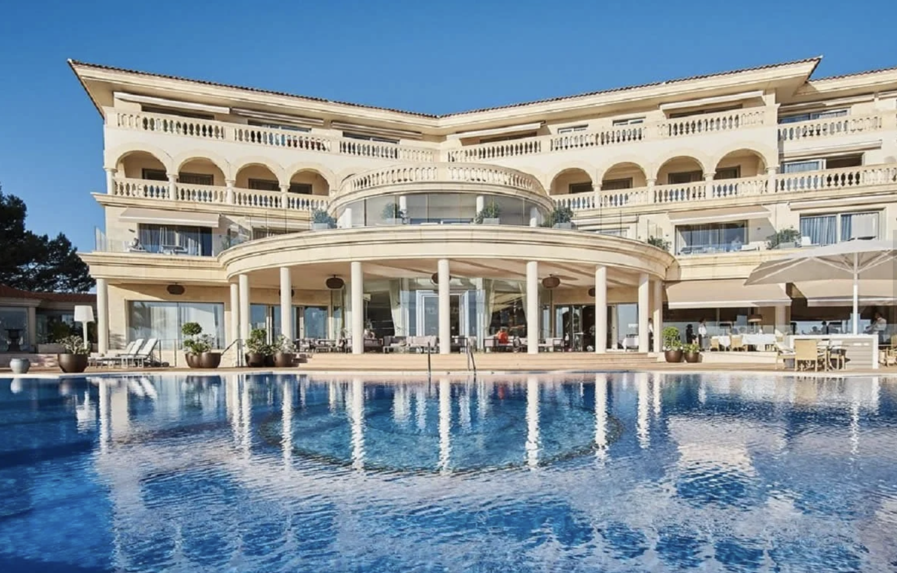 Edificio lujoso con varias terrazas y balustradas, frente a una piscina con agua azul y reflejos del edificio, en un día soleado con cielo despejado.